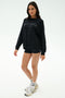 A woman stands in a studio wearing the SPLITS59 Andie Oversized Fleece Varsity Graphic Sweatshirt in black, black shorts, white sneakers, and white socks.
