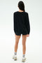 A person faces away, wearing the SPLITS59 Andie Oversized Fleece Varsity Graphic Sweatshirt in black with shorts, white socks with black stripes, and white sneakers, set against a plain light background.