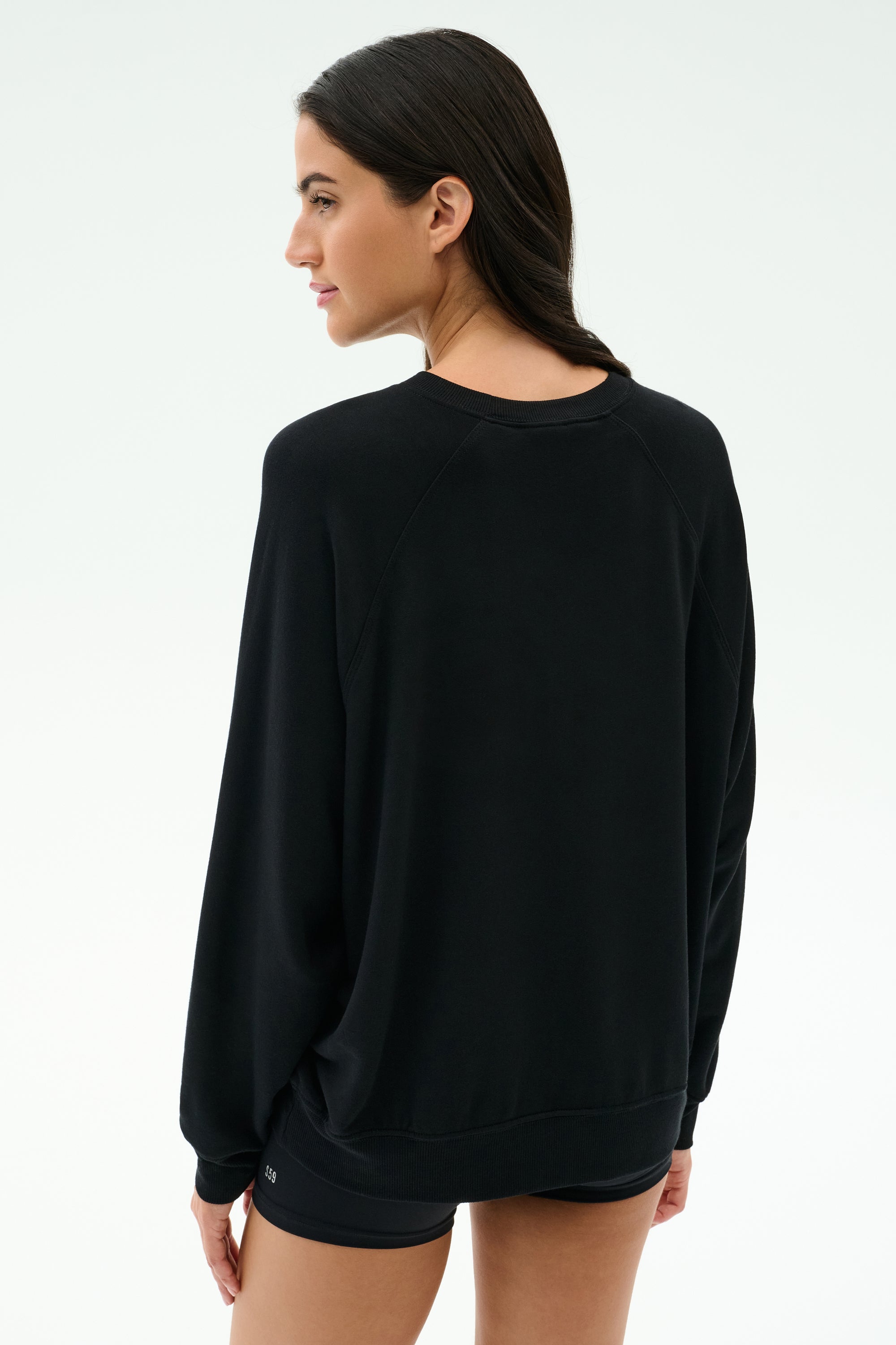 A woman stands against a light background, facing away and looking over her shoulder, wearing the SPLITS59 Andie Oversized Fleece Varsity Graphic Sweatshirt in black by Splits59 Los Angeles.