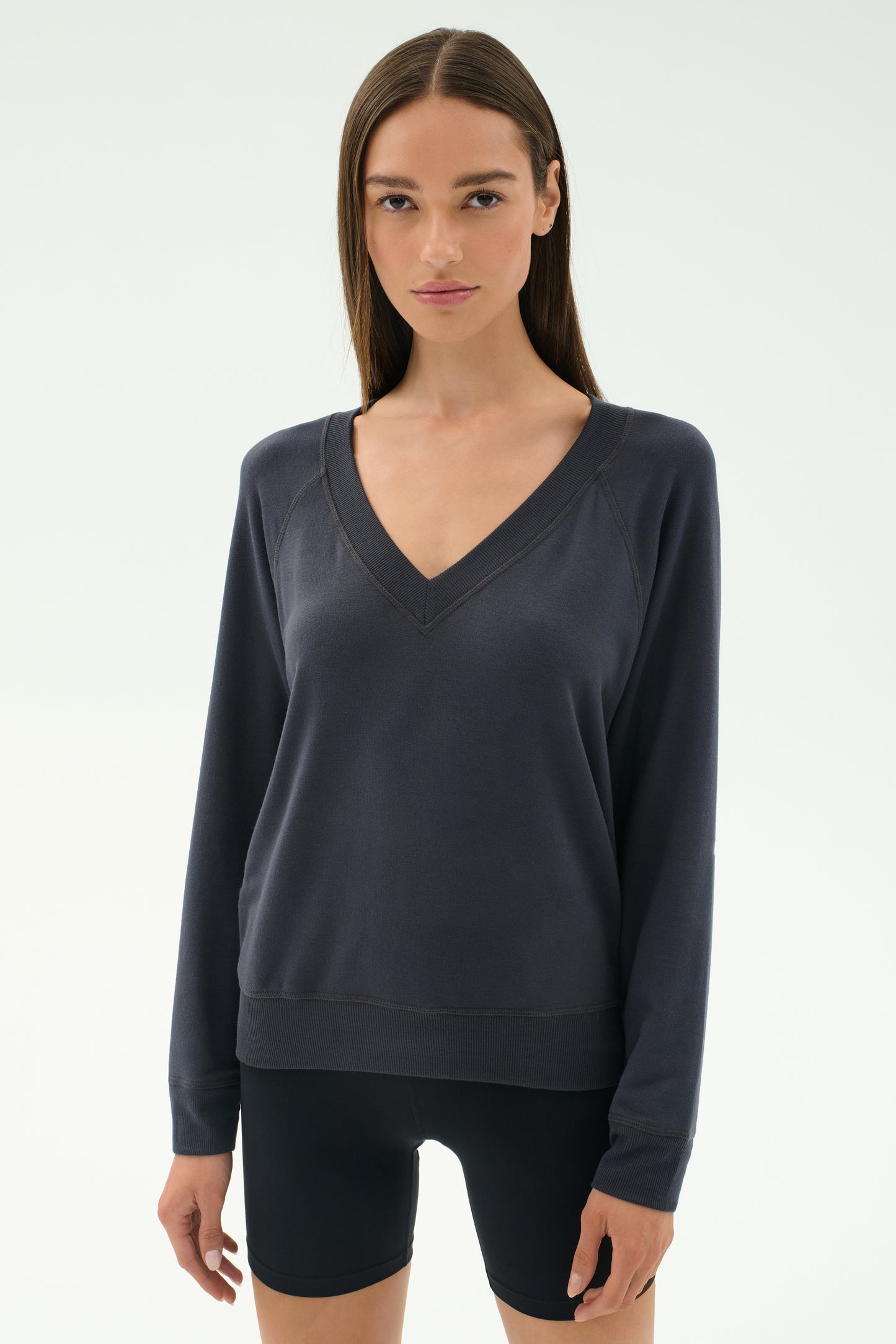 A woman stands against a light background, wearing the SPLITS59 Bennie Fleece V Neck Sweatshirt in Graphite and black shorts. This chic pullover is crafted in Los Angeles.