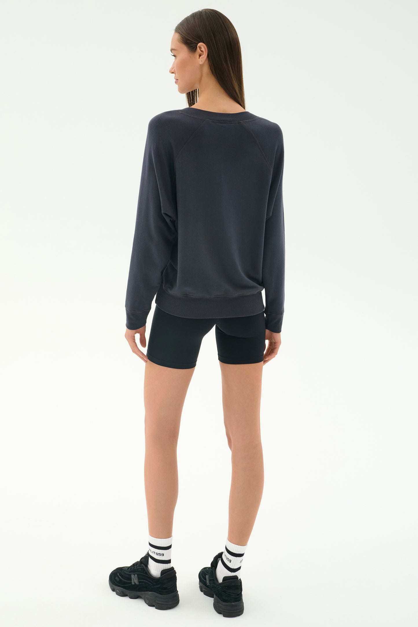 A woman faces away, wearing the SPLITS59 Bennie Fleece V-Neck Sweatshirt in Graphite with black cycling shorts, white socks, and black sneakers—styled simply against a plain white background.