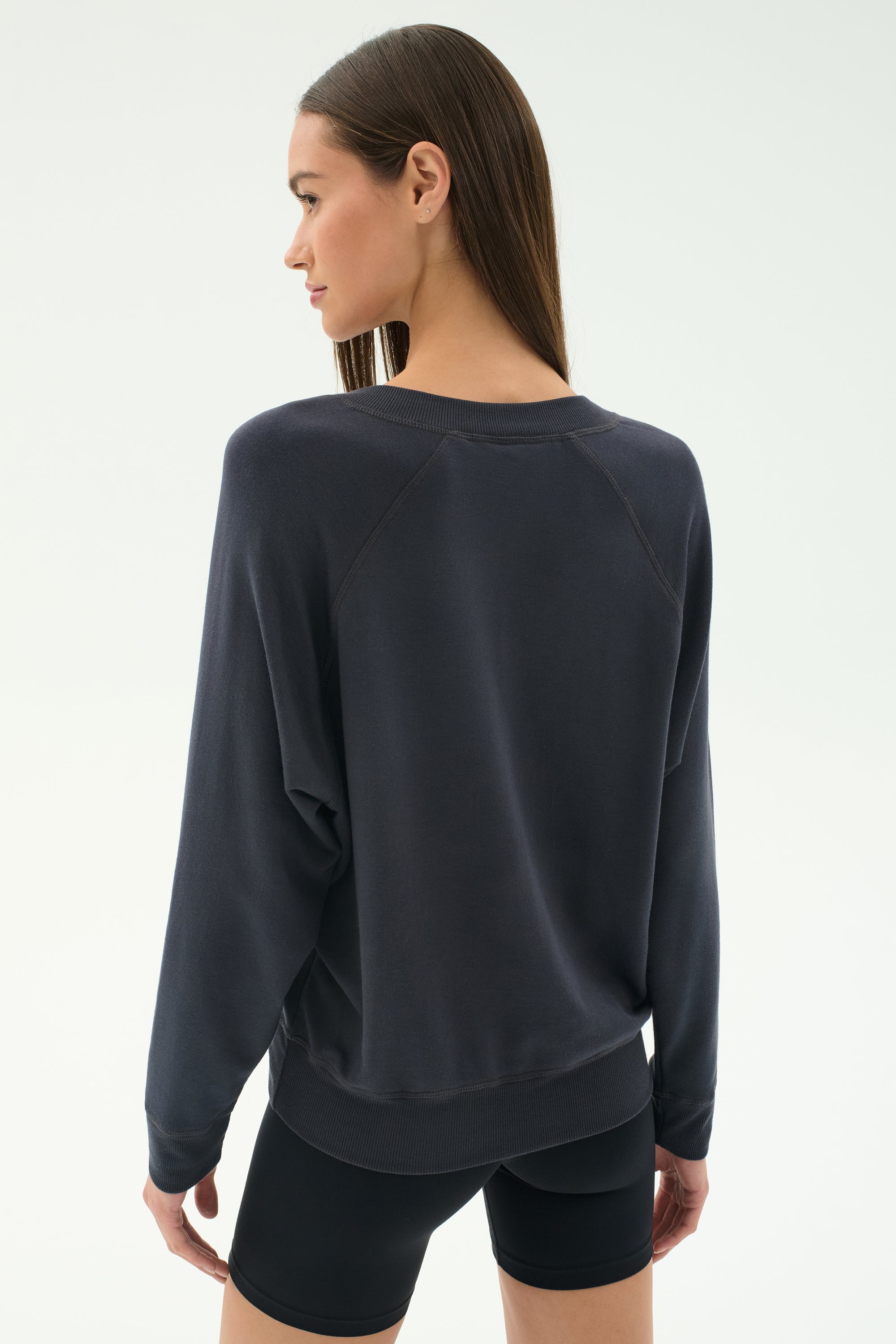 A woman with long brown hair wears the SPLITS59 Bennie Fleece V-Neck Sweatshirt in Graphite and black shorts, standing with her back to the camera in a minimal, light-toned setting.