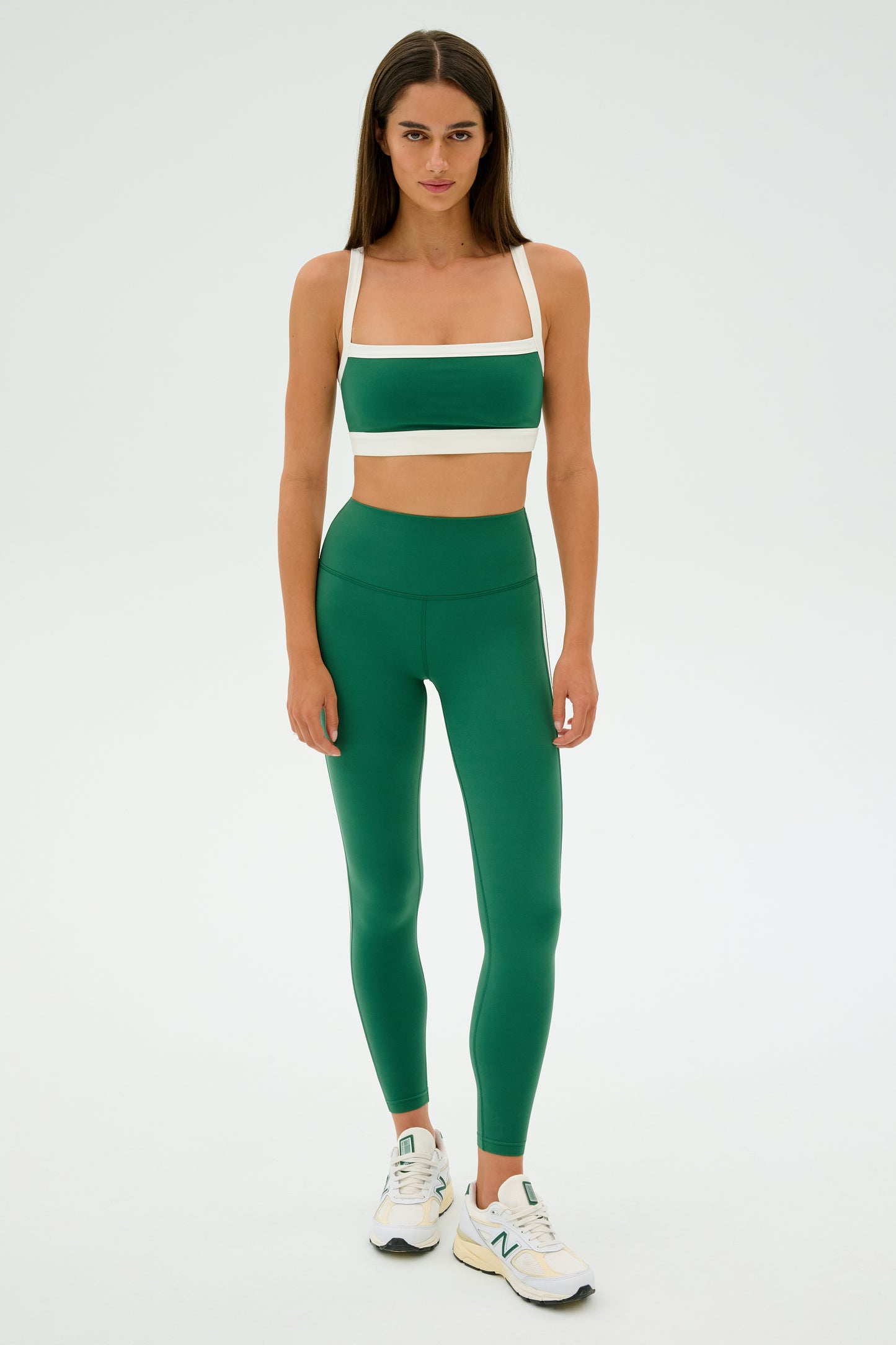 Someone exudes confidence in a green and white athletic outfit with SPLITS59 Clare High Waist Rigor 7/8 leggings, paired with white sneakers against a plain backdrop.