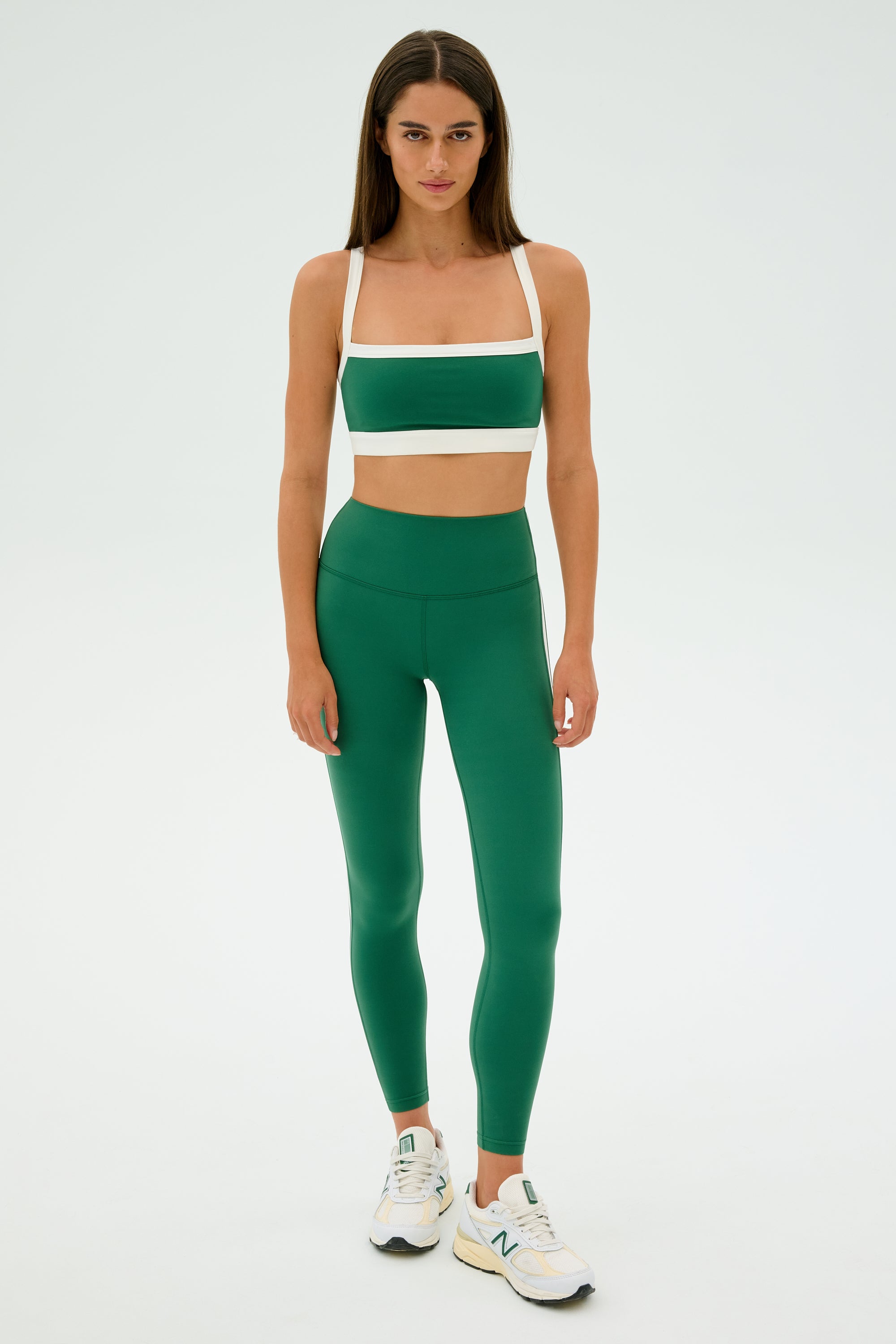 Someone exudes confidence in a green and white athletic outfit with SPLITS59 Clare High Waist Rigor 7/8 leggings, paired with white sneakers against a plain backdrop.