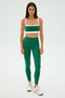 Someone exudes confidence in a green and white athletic outfit with SPLITS59 Clare High Waist Rigor 7/8 leggings, paired with white sneakers against a plain backdrop.