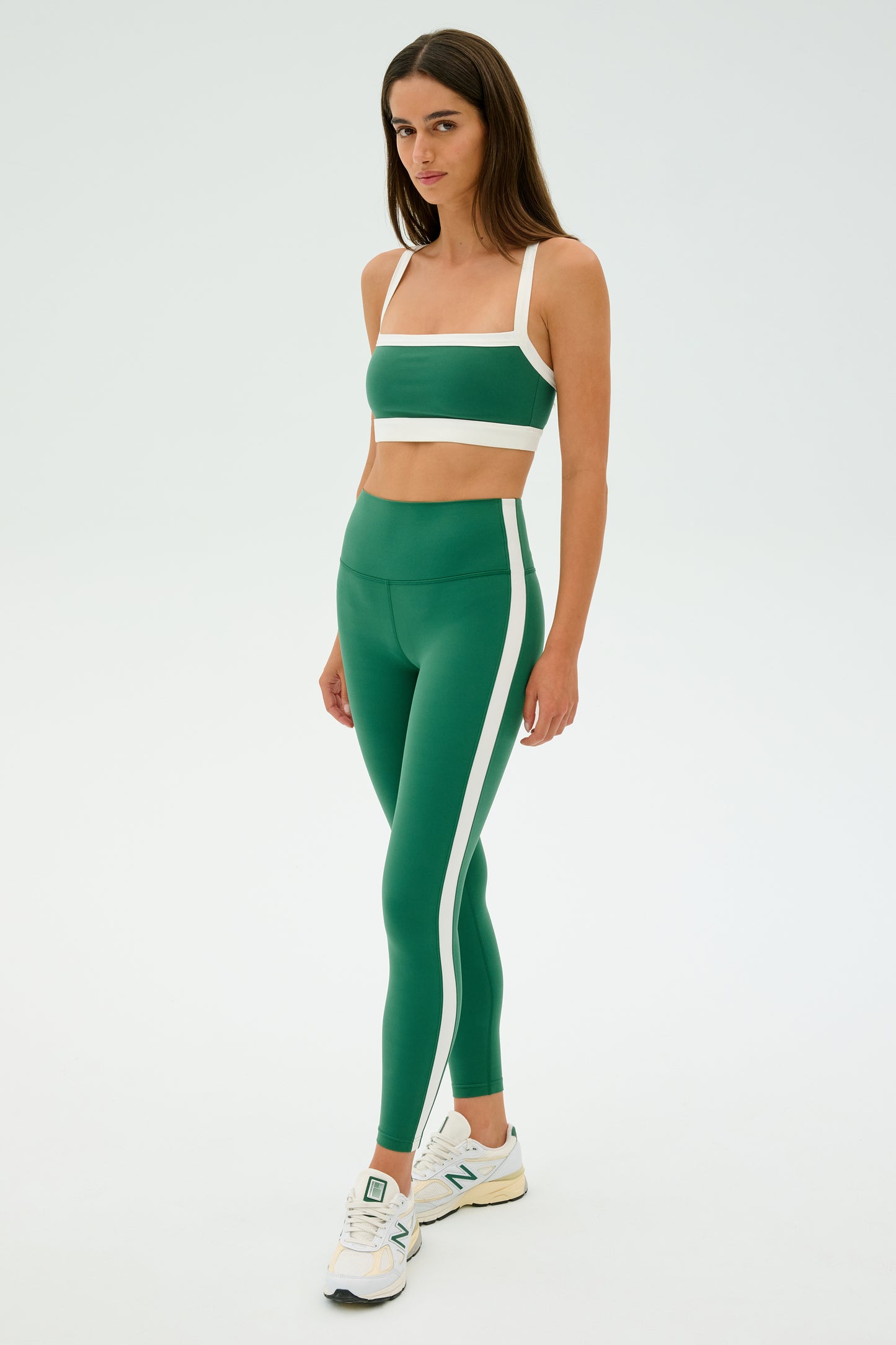 A person in an SPLITS59 Clare High Waist Rigor 7/8 Leggings in Arugula/White, green sports bra, and white sneakers stands against a plain background.