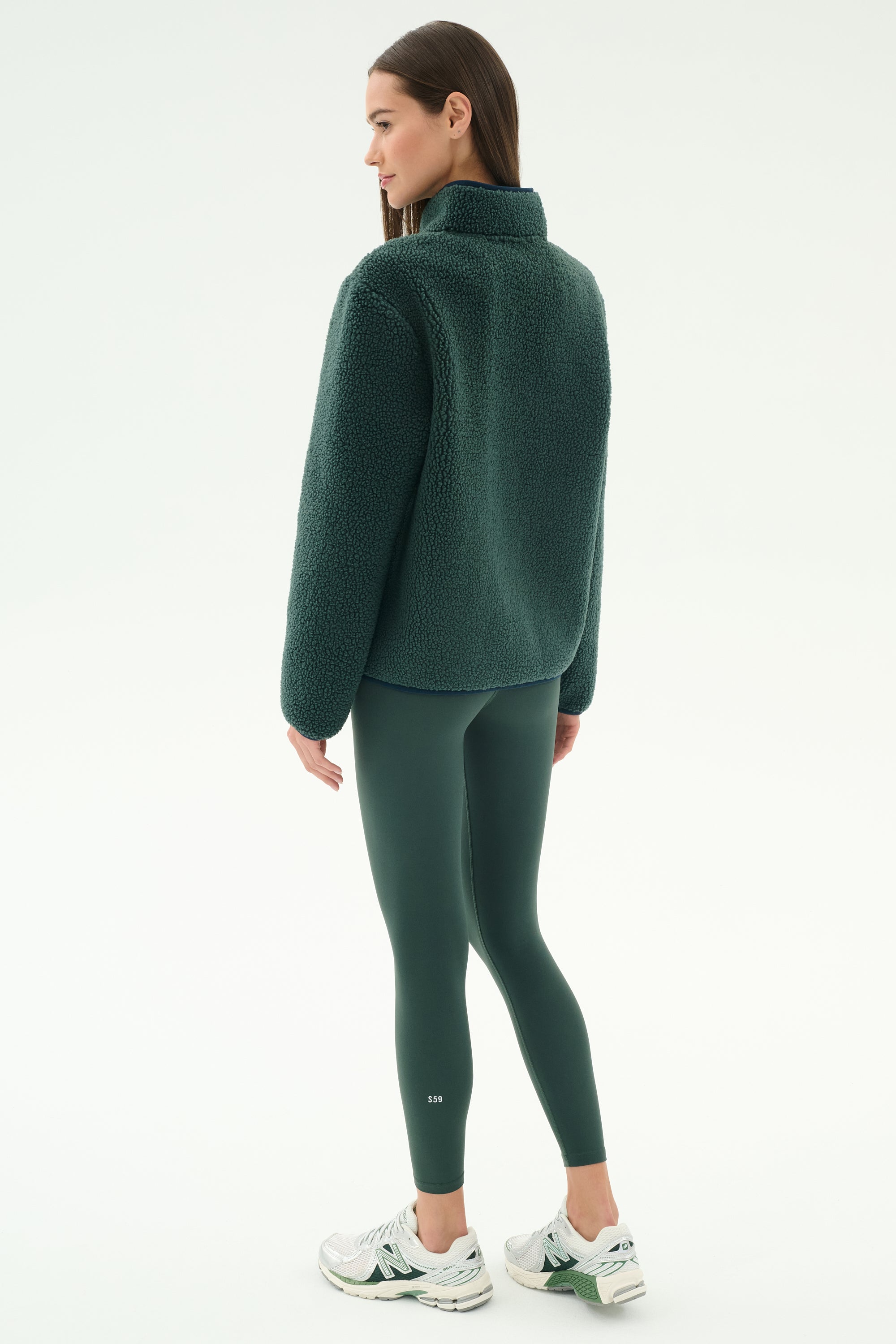 A woman faces away, wearing the SPLITS59 Harris Sherpa Jacket in Military/Indigo, with matching leggings and white sneakers against a plain white background.