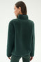 A woman stands with her back turned, wearing the SPLITS59 Harris Sherpa Jacket in Military/Indigo and green leggings against a plain light background.