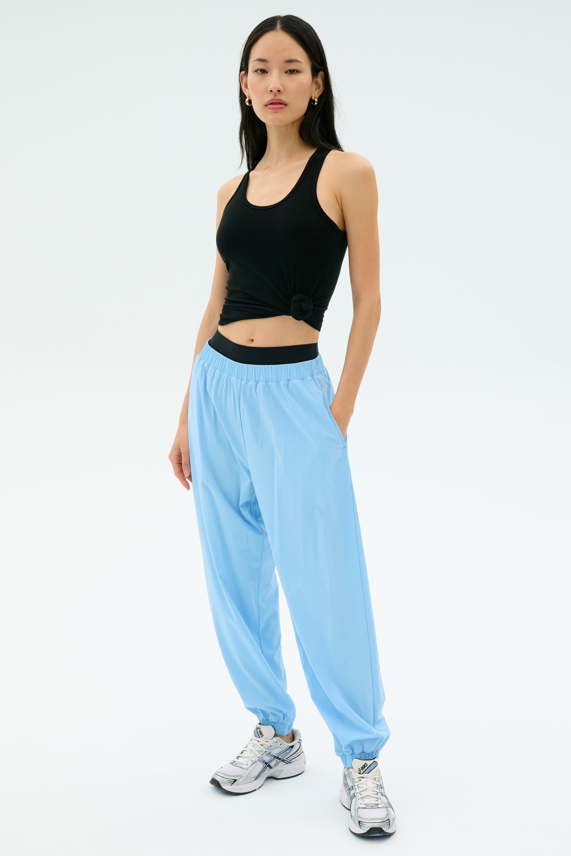 A person wearing a black sleeveless top, SPLITS59 Megan Woven Nylon Jogger in Skylight, and white sneakers stands against a plain light background.