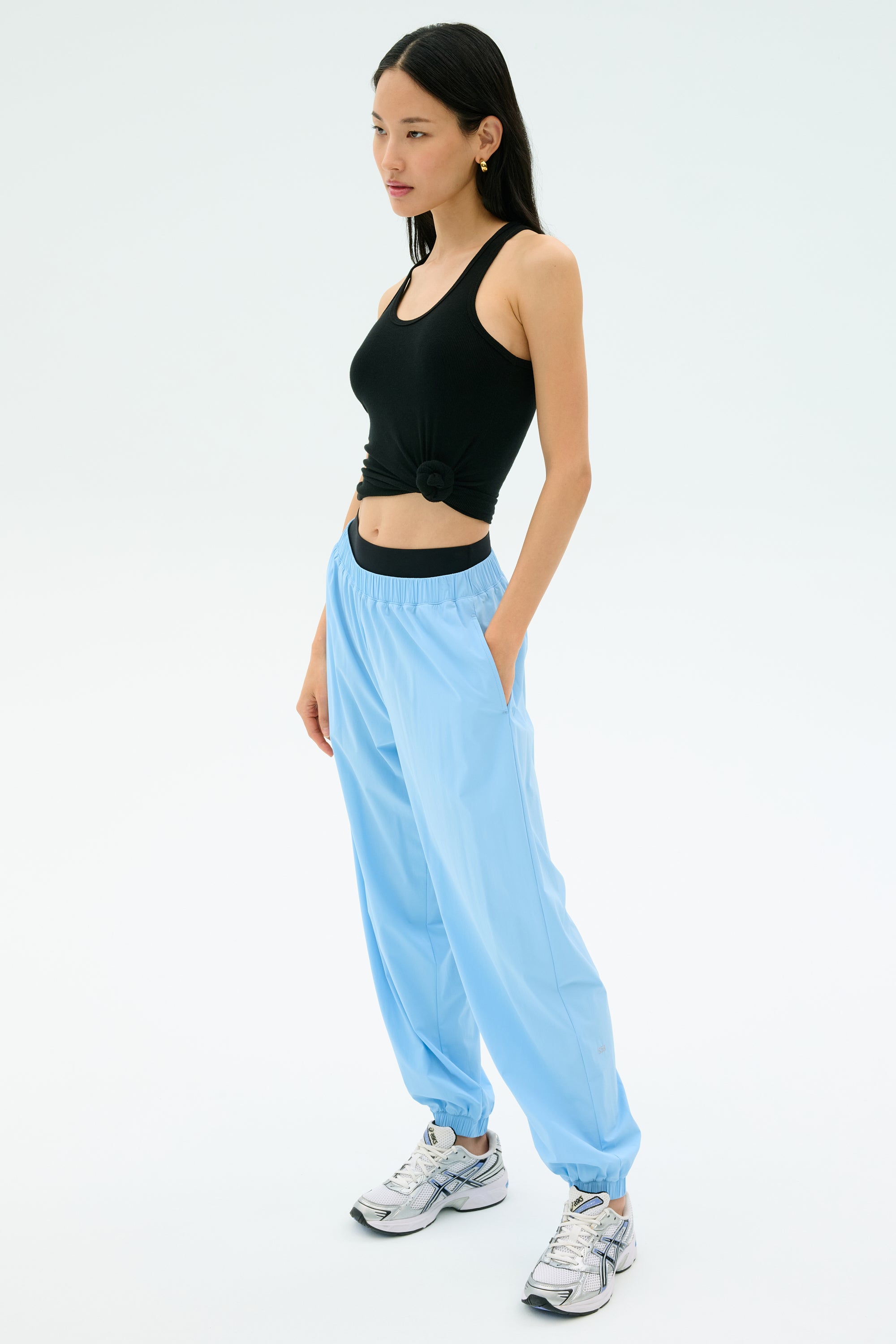 A woman stands against a plain background wearing SPLITS59's Megan Woven Nylon Jogger in Skylight, paired with a black sleeveless crop top and white sneakers, hands in her pockets.