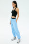 A woman stands against a plain background wearing SPLITS59's Megan Woven Nylon Jogger in Skylight, paired with a black sleeveless crop top and white sneakers, hands in her pockets.