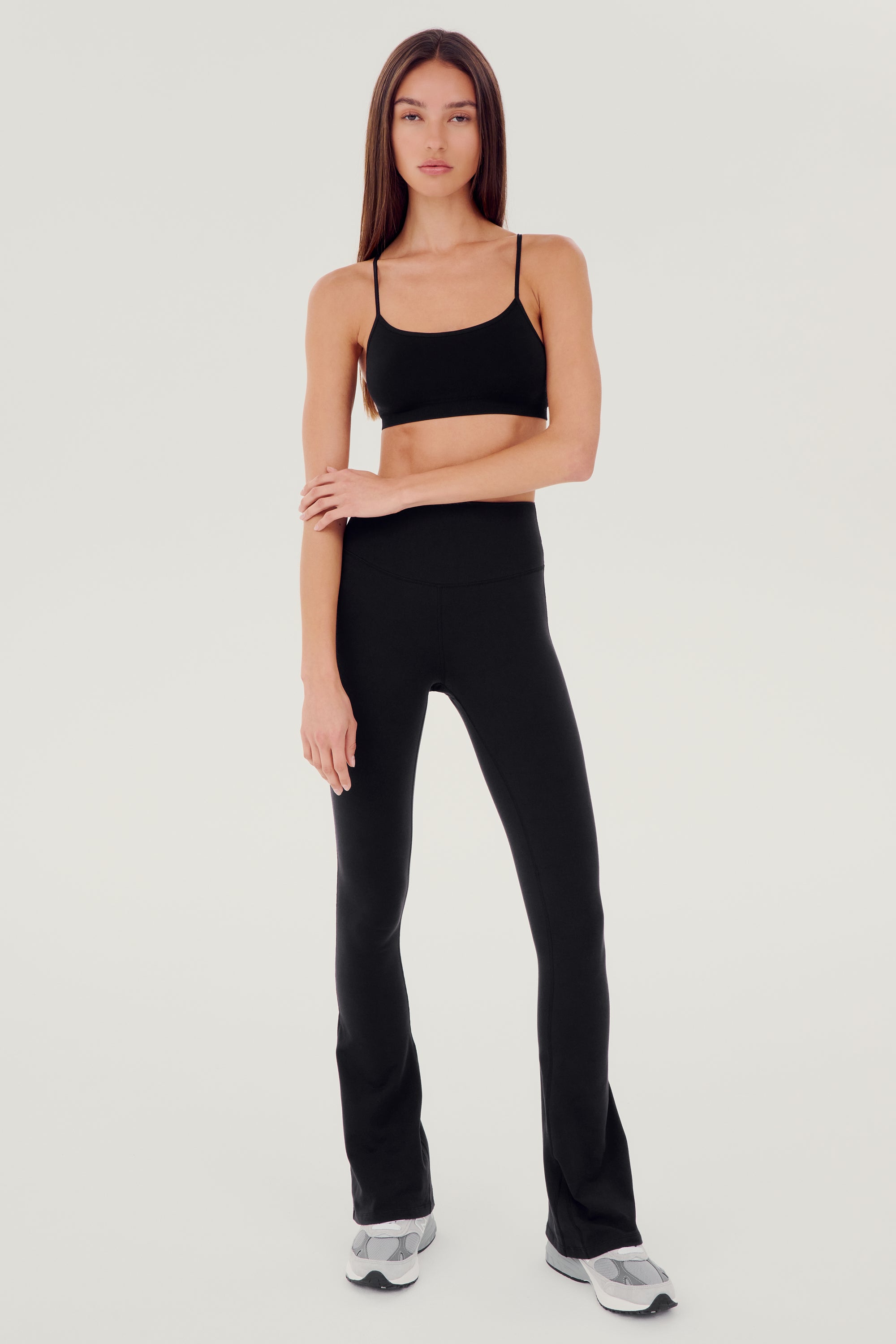 Full front view of woman with dark straight hair wearing black high waist below ankle length legging with wide flared bottoms and black racerback bra with spaghetti straps. Paired with white and gray shoes.