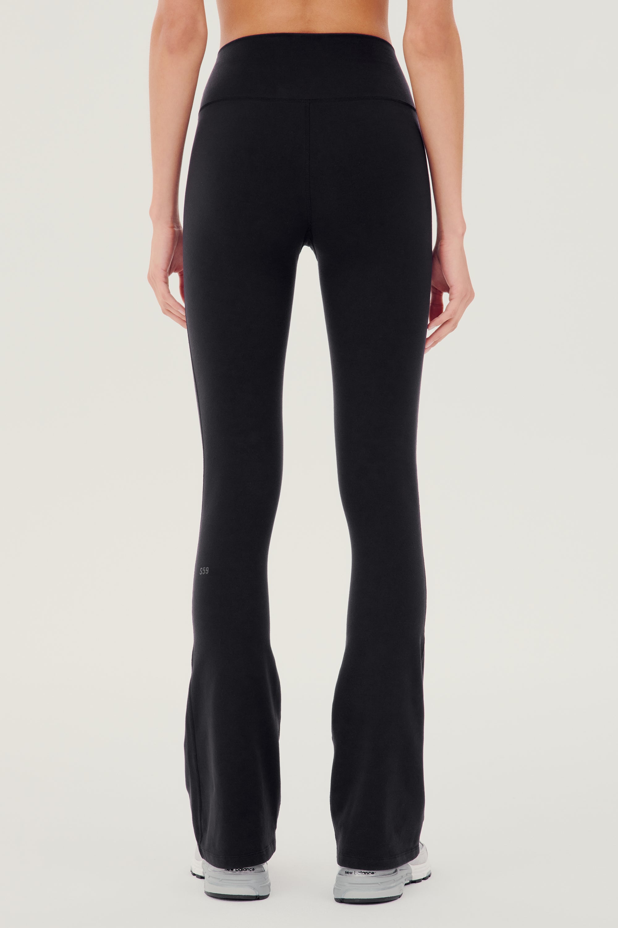 Back side view of woman wearing black high waist below ankle length legging with wide flared bottoms and gray S59 logo on bottom left calf. Paired with white and gray shoes.