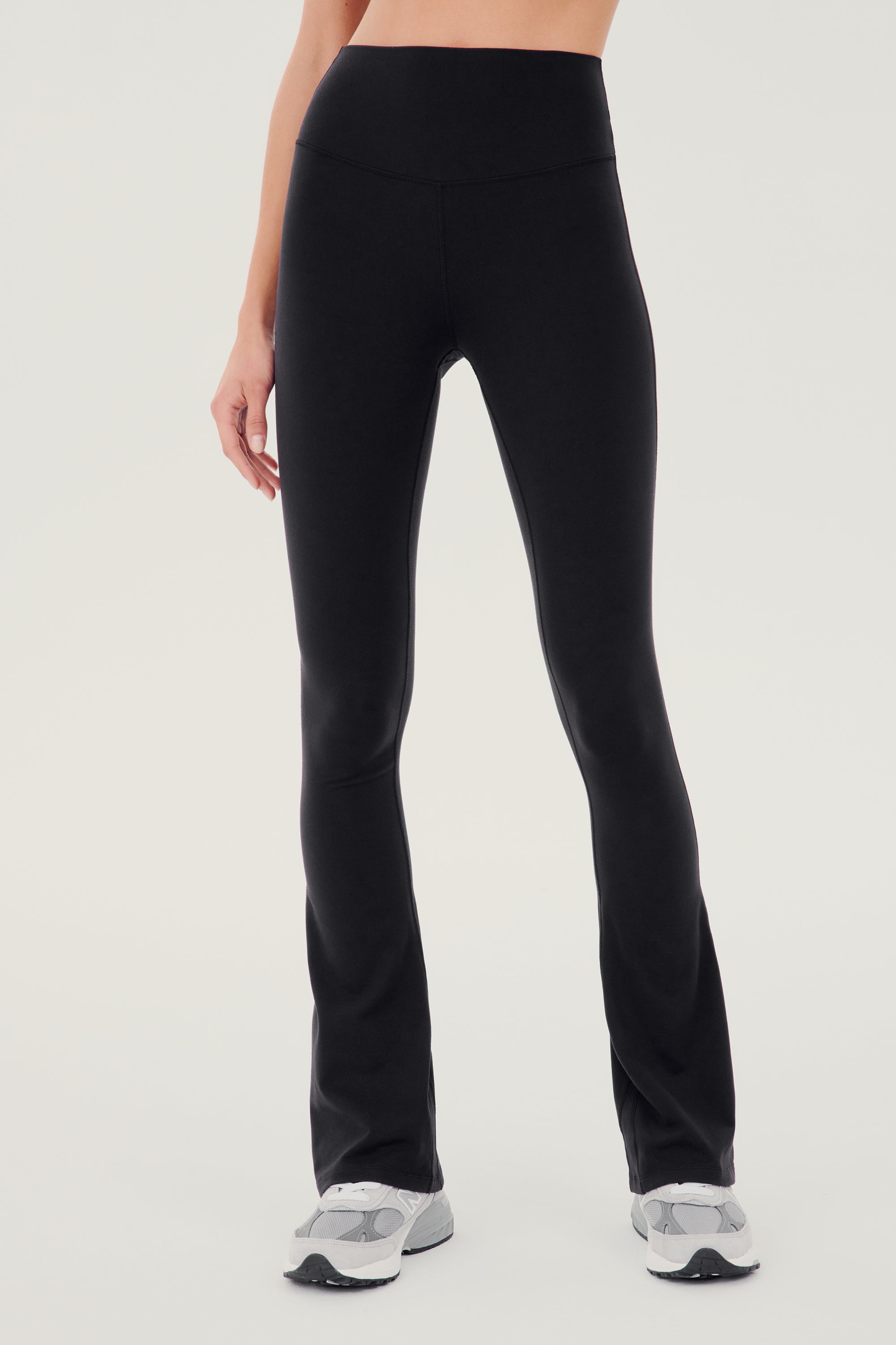 Front view of woman wearing black high waist below ankle length legging with wide flared bottoms. Paired with white and gray shoes.