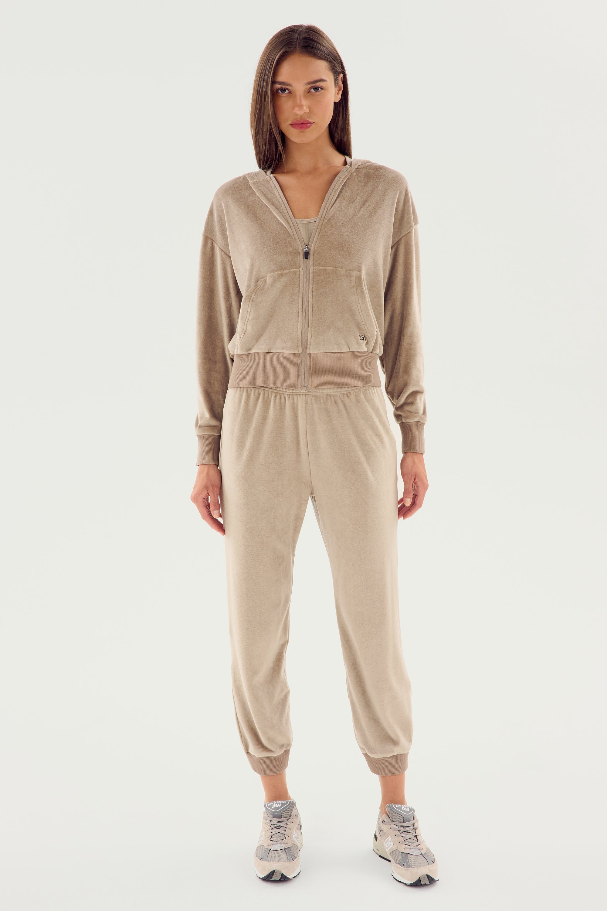 A person stands against a plain background wearing a SPLITS59 Rhys Velour Jogger - Latte tracksuit consisting of a zip-up jacket and jogger pants, paired with white sneakers. This casual wear outfit is perfect for lounging or running errands in style.