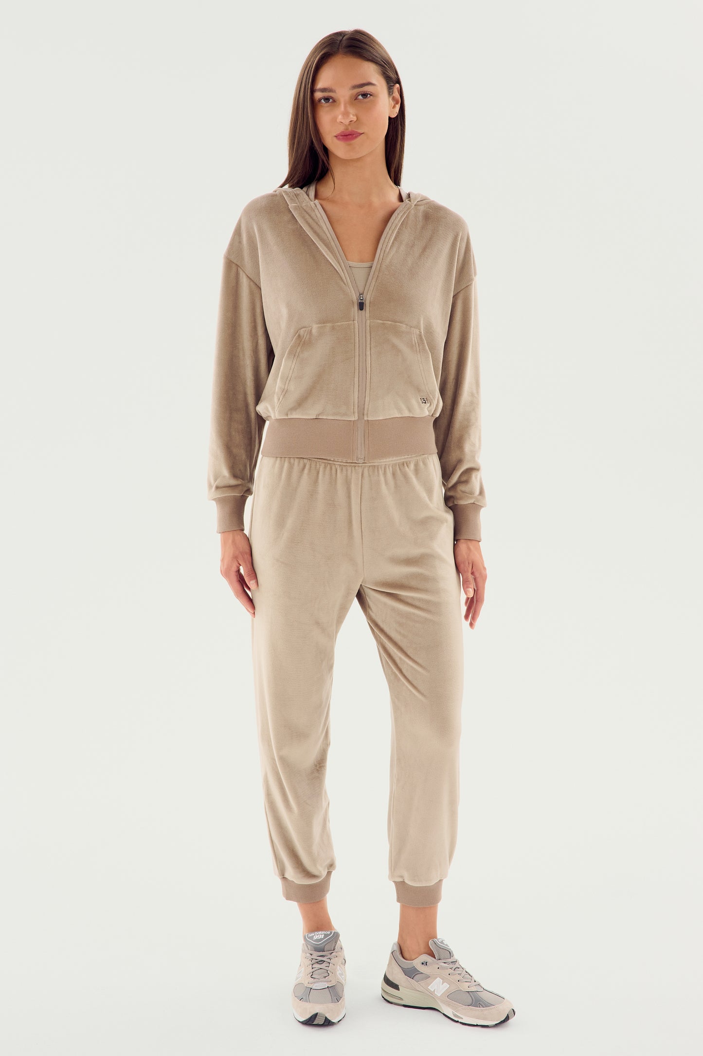 A woman stands facing forward, wearing the SPLITS59 Rhys Velour Zip Up Hoodie - Latte with matching joggers, paired with white sneakers. The ensemble exudes comfort and versatility against the plain white background.