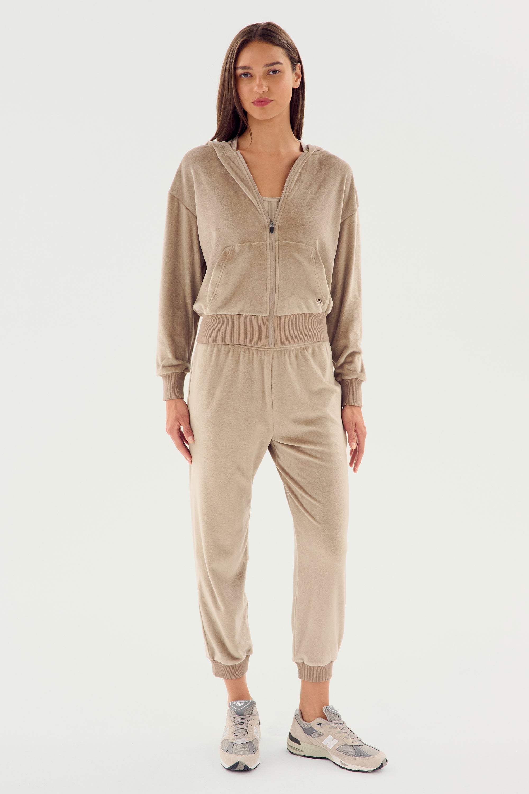 A woman stands facing forward, wearing the SPLITS59 Rhys Velour Zip Up Hoodie - Latte with matching joggers, paired with white sneakers. The ensemble exudes comfort and versatility against the plain white background.