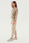 A woman stands in profile wearing a beige tracksuit with a hoodie and SPLITS59 Rhys Velour Jogger - Latte pants, paired with light-colored sneakers, embodying casual wear against a plain white background.