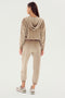 A woman is standing with her back to the camera, wearing a beige SPLITS59 Rhys Velour Jogger - Latte tracksuit and sneakers. Her long hair cascades down her back, and the tracksuit has a hood resting on her shoulders, exuding comfort and casual wear vibes.