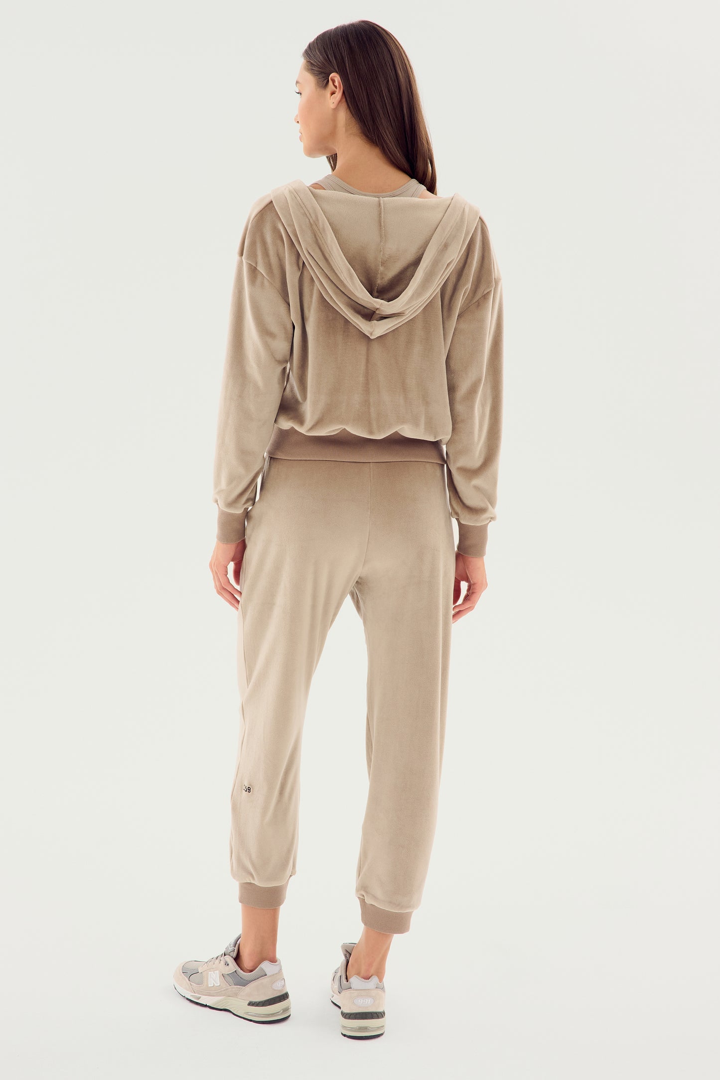 A person is standing with their back to the camera, wearing a beige velour tracksuit featuring the SPLITS59 Rhys Velour Zip Up Hoodie - Latte and matching pants, along with gray athletic shoes. The ensemble exudes comfort and versatility.