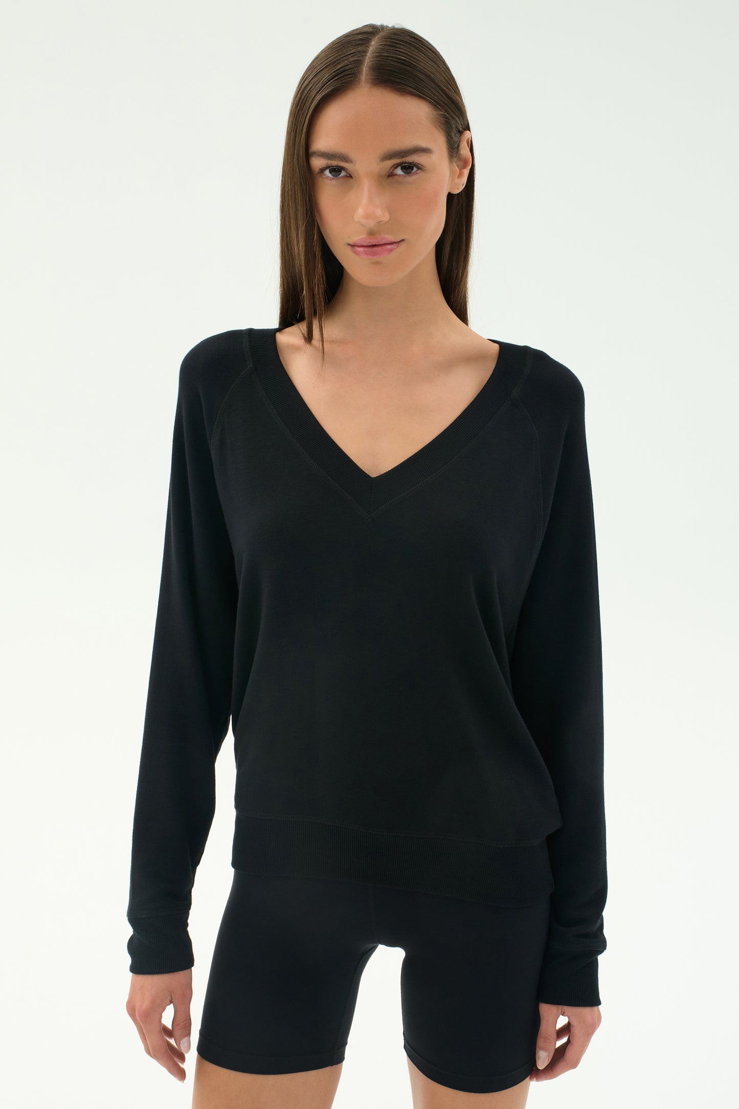 A woman with straight brown hair models the SPLITS59 Bennie Fleece V-Neck Sweatshirt in black and fitted shorts against a white background. This stylish look is made in Los Angeles.