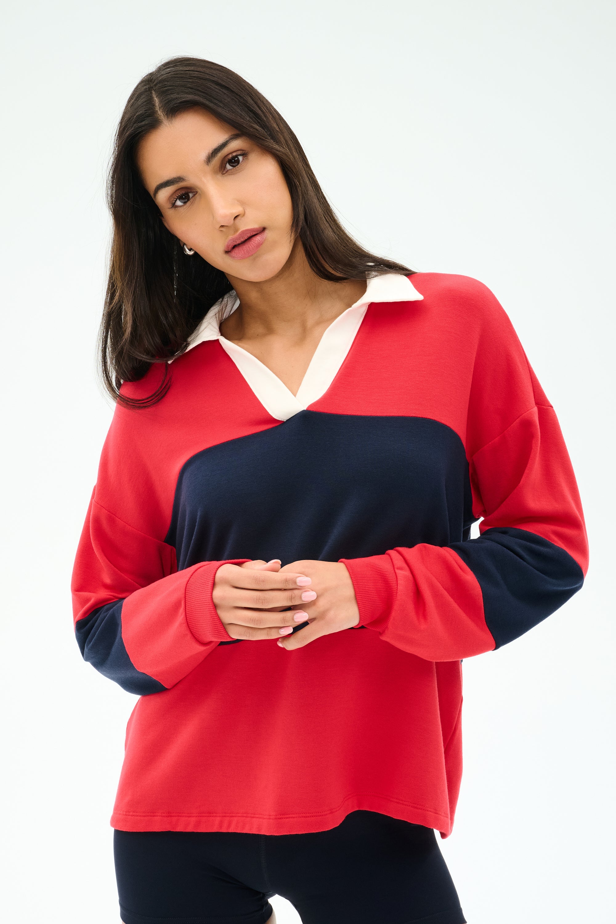 A woman with long brown hair wears the SPLITS59 Katie Fleece Rugby in Pirate Red/Indigo, featuring a white collar, standing against a plain light background.