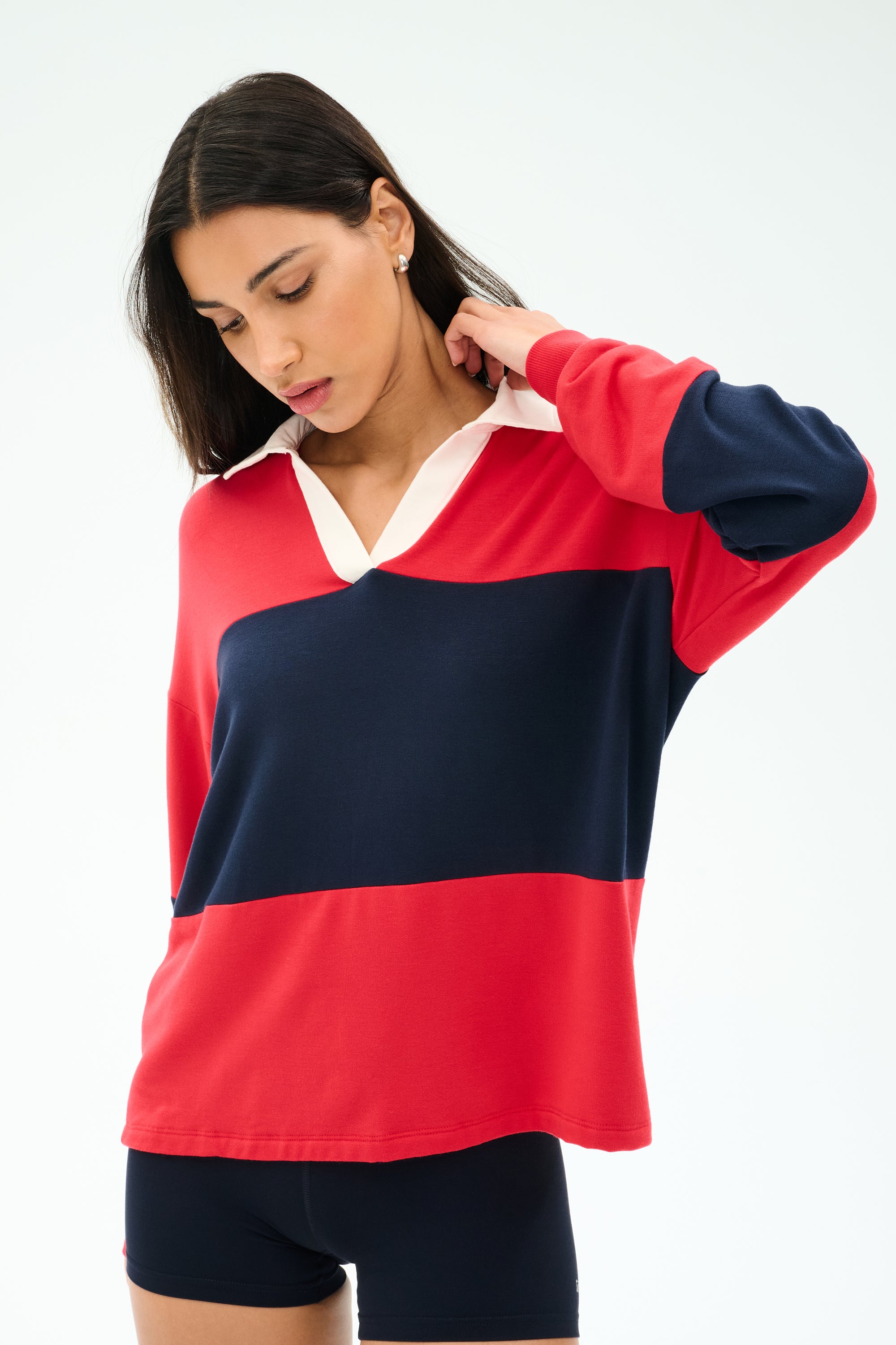 A person wears the SPLITS59 Katie Fleece Rugby in Pirate Red/Indigo with navy shorts, standing against a white background and looking down while touching their neck.