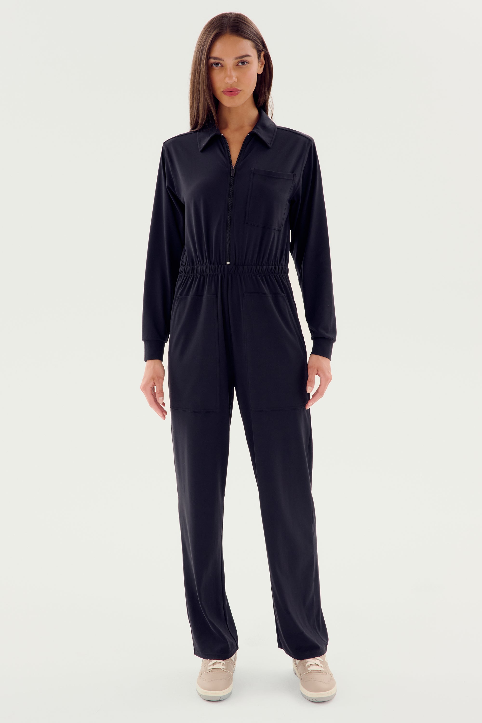A person wearing a stylish SPLITS59 Ollie Airweight Boiler Suit - Black with beige shoes stands against a plain white backdrop, showcasing a sleek modern style.
