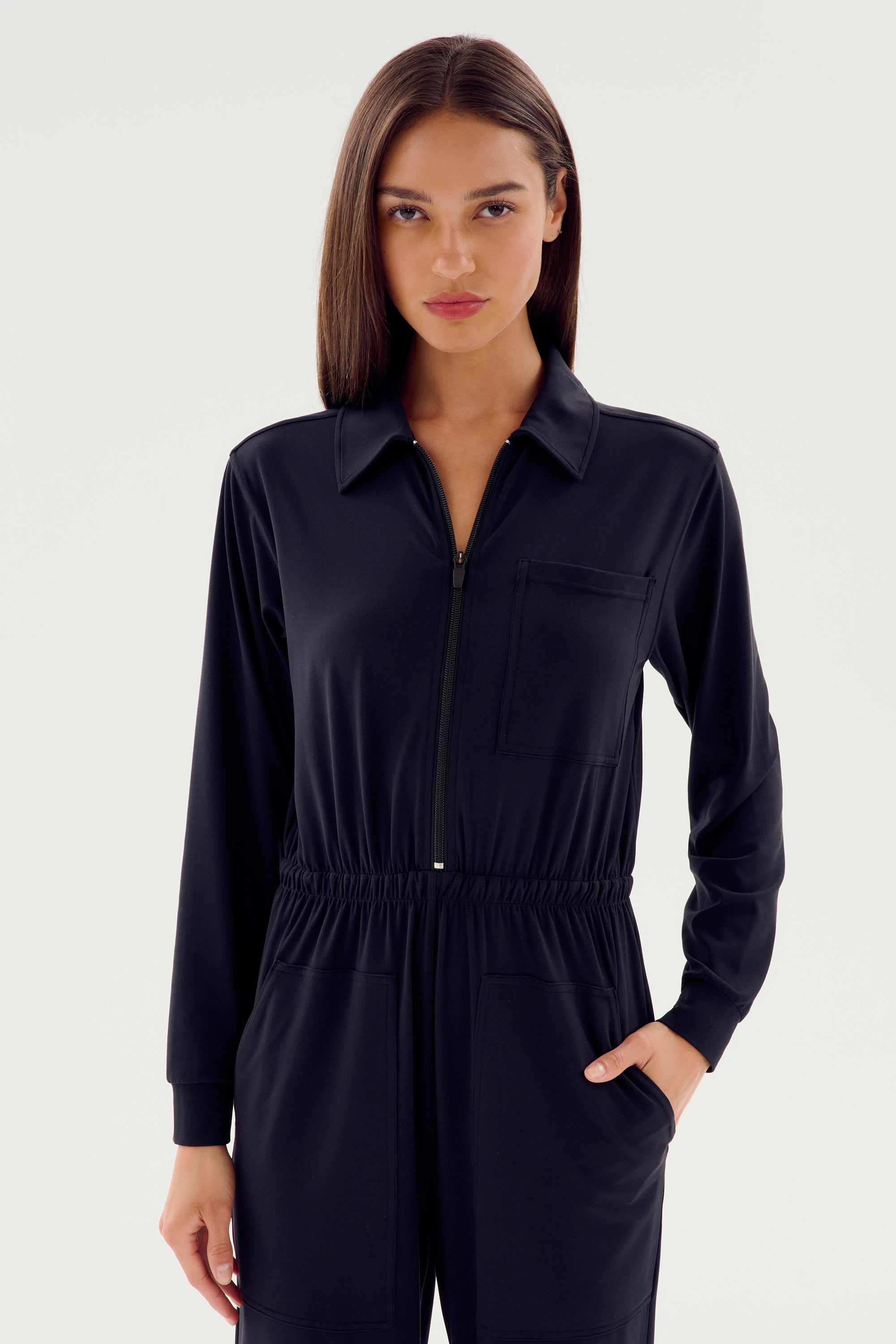 A person wearing the luxurious SPLITS59 Ollie Airweight Boiler Suit in black, featuring a zipper and pockets, stands against a plain backdrop.