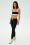Dressed in a black and white sports bra, SPLITS59 Sonja Fleece Sweatpant - Black, and silver sneakers, this person stands against a plain background—ideal for cool weather workouts.
