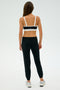 A woman facing away, wearing a black and white sports bra, Sonja Fleece Sweatpant in Black by SPLITS59, and silver sneakers, stands against a plain white background.