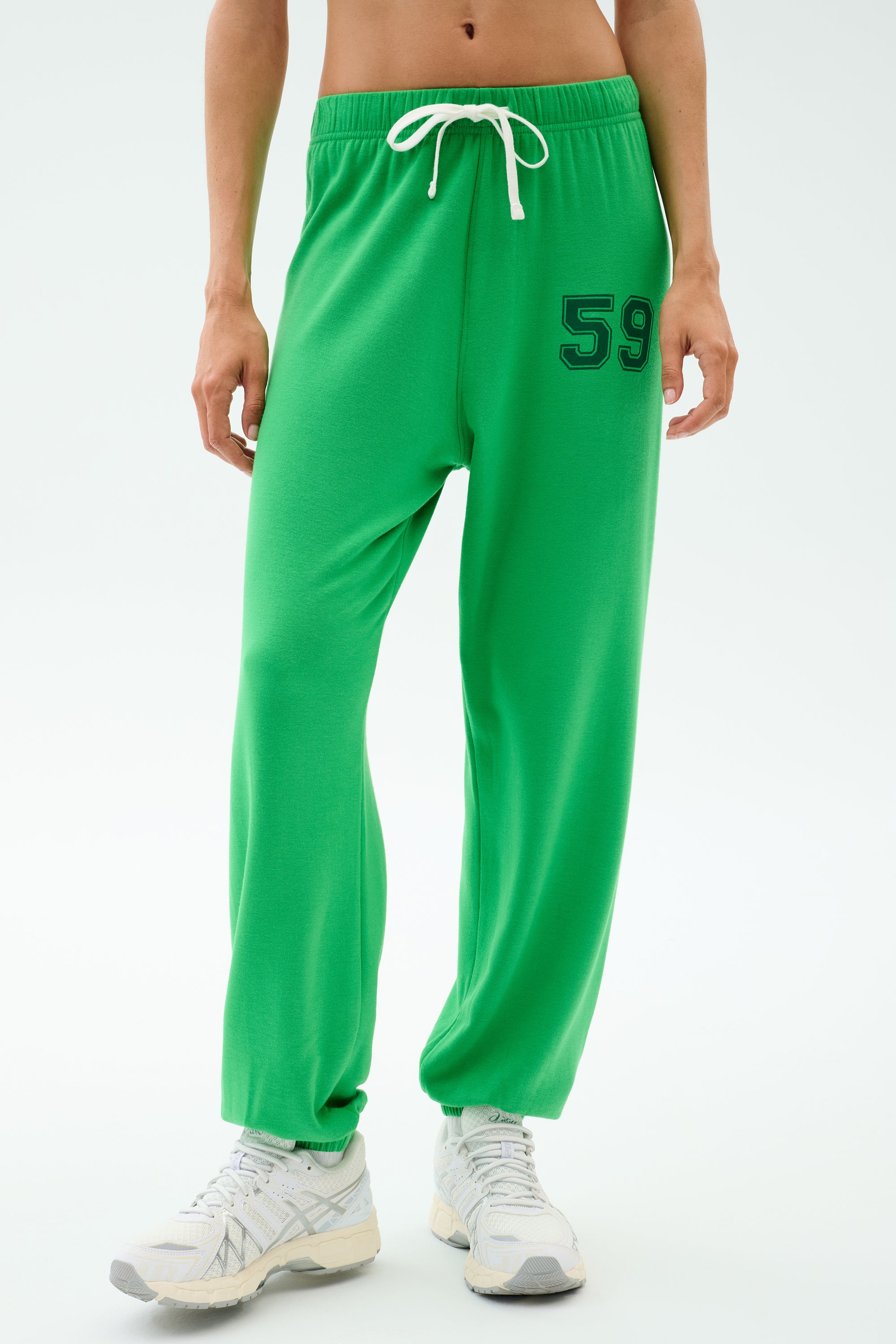 Wearing SPLITS59’s Andie Oversized Fleece Varsity 59 Sweatpant in Grass/59 with a white drawstring and "59" on the left thigh, paired with white sneakers—ideal for warm weather workouts against a plain backdrop.
