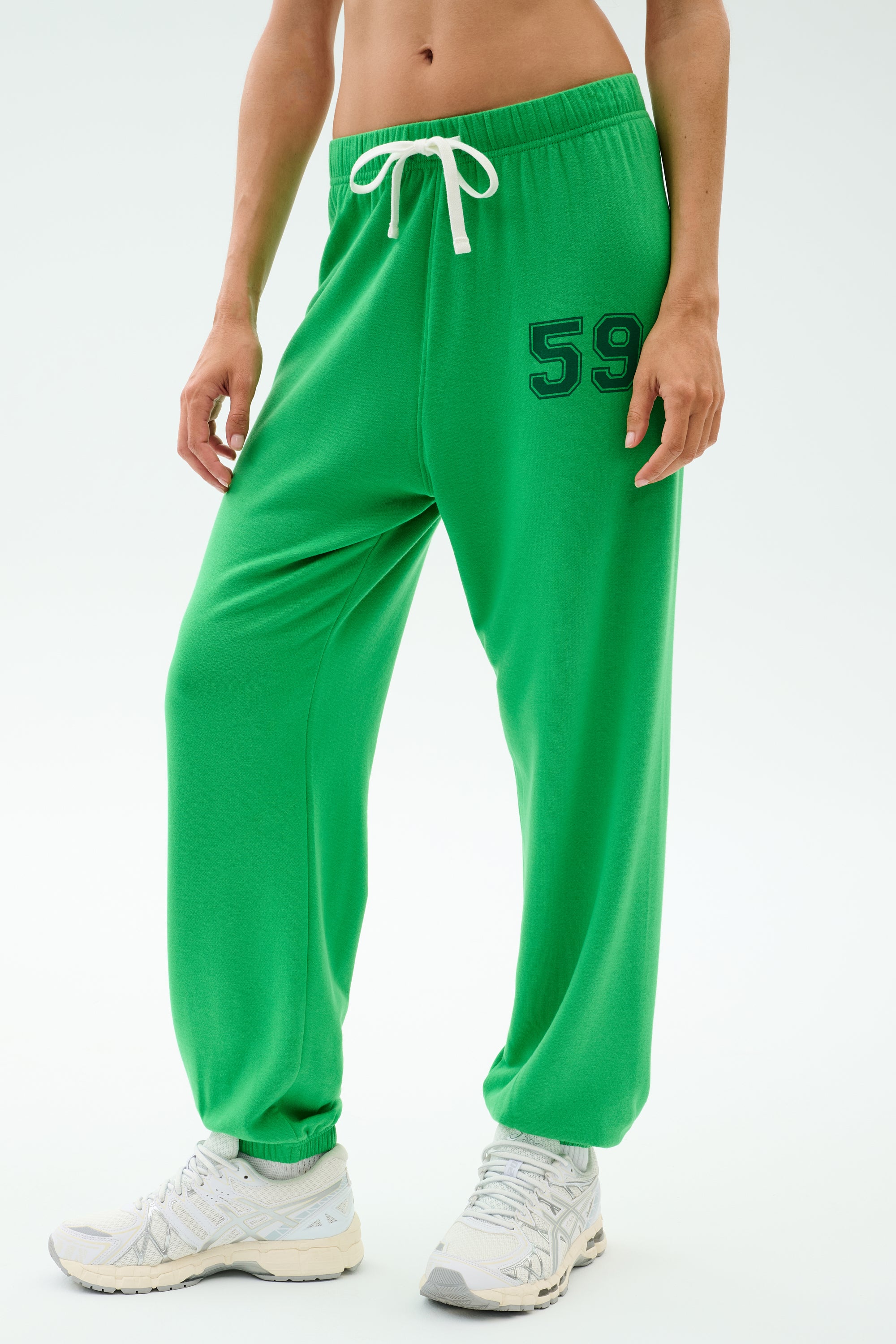 A person wears the SPLITS59 Andie Oversized Fleece Varsity 59 Sweatpant in Grass/59, featuring a bright green color, white drawstring, and number 59 on the left thigh with white athletic shoes against a plain background.