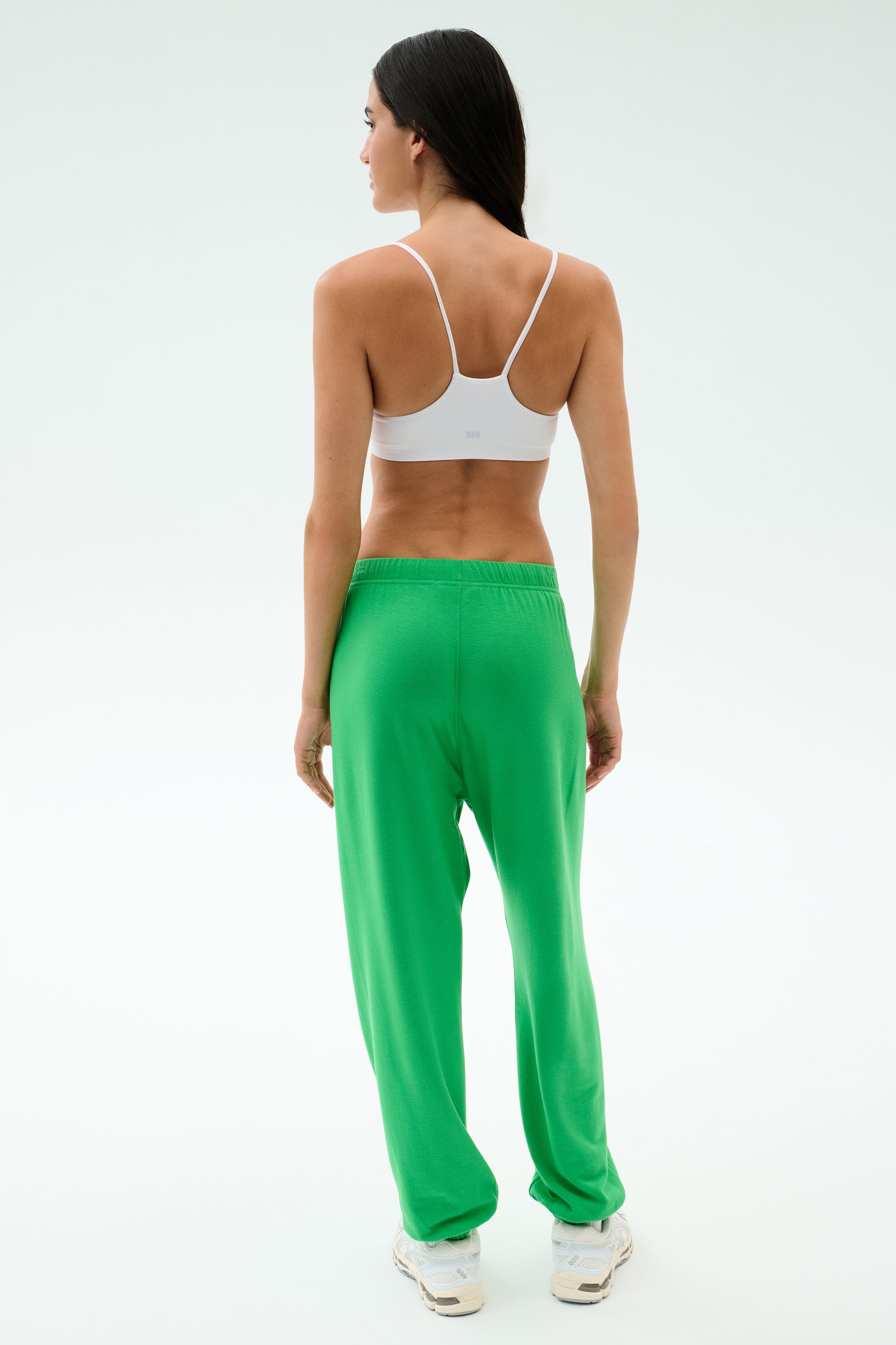 A woman faces away, wearing a white sports bra, white sneakers, and the SPLITS59 Andie Oversized Fleece Varsity 59 Sweatpant in Grass/59 against a plain light background—ideal for warm weather workouts.