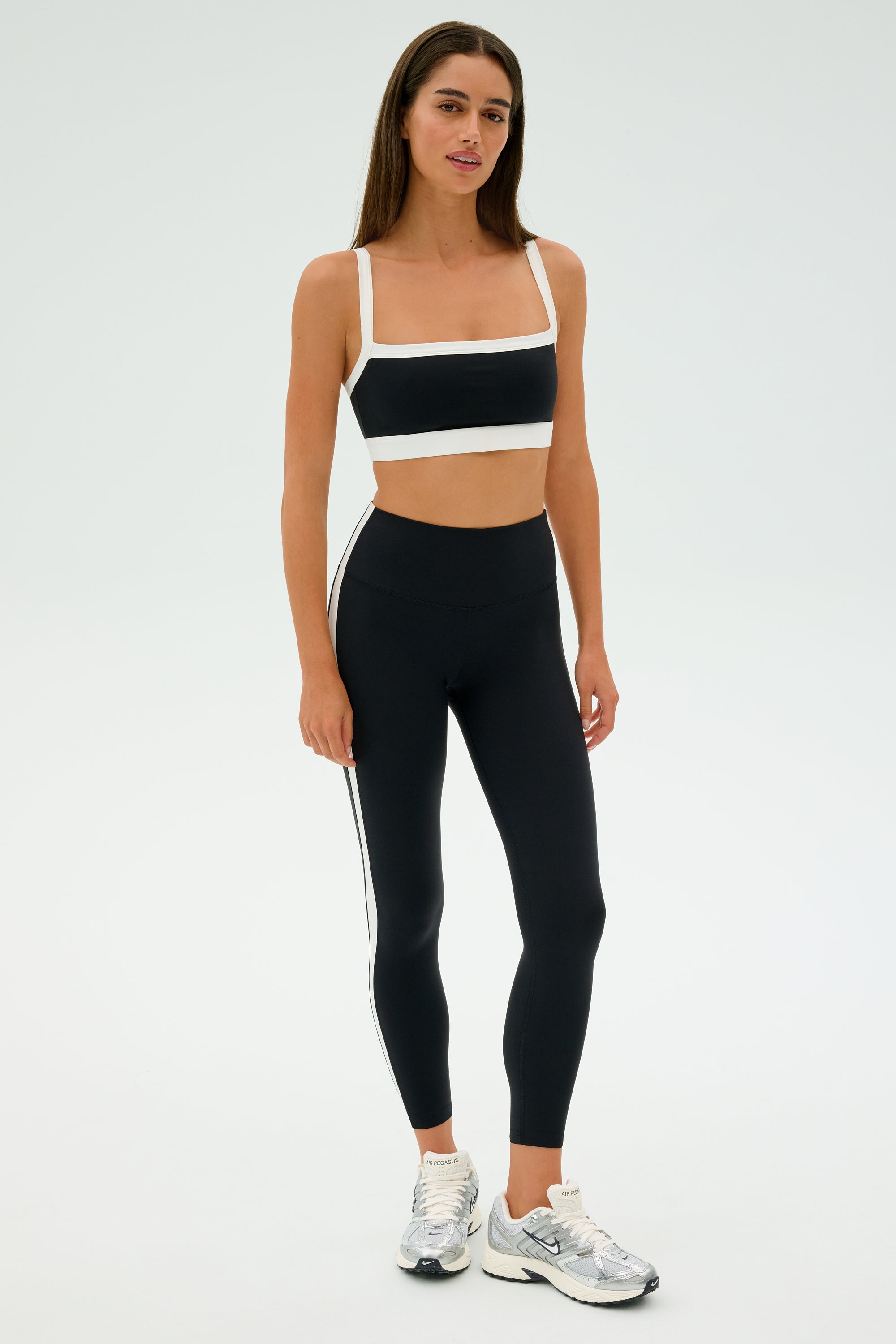A woman is wearing SPLITS59's Clare High Waist Rigor 7/8 leggings in black and white, a matching sports bra, and silver-white athletic shoes against a plain background.