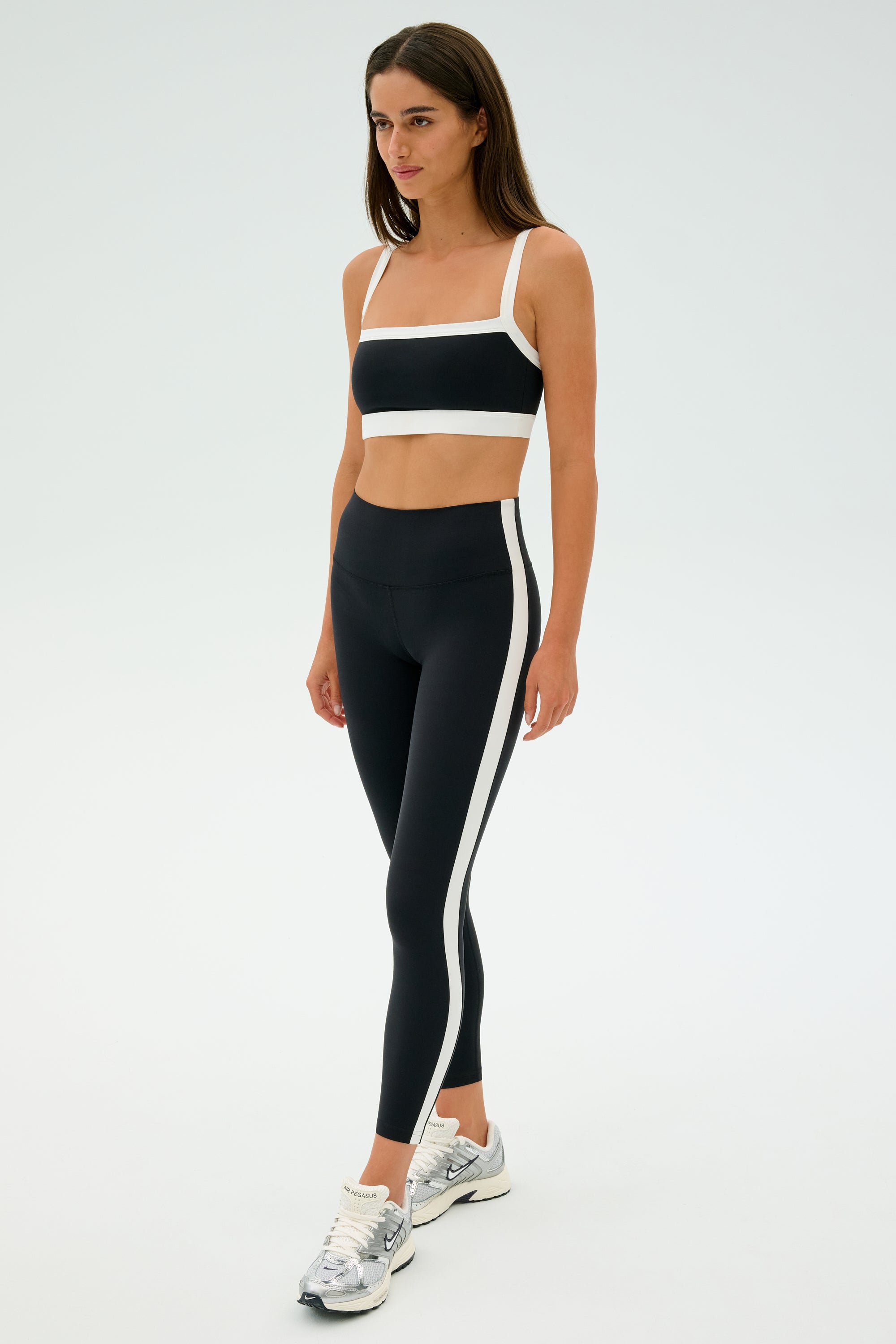 Against a plain background, a woman wears SPLITS59's Clare High Waist Rigor 7/8 leggings in Black/White with a cropped top and silver sneakers, all made from high-performance Rigor fabric.