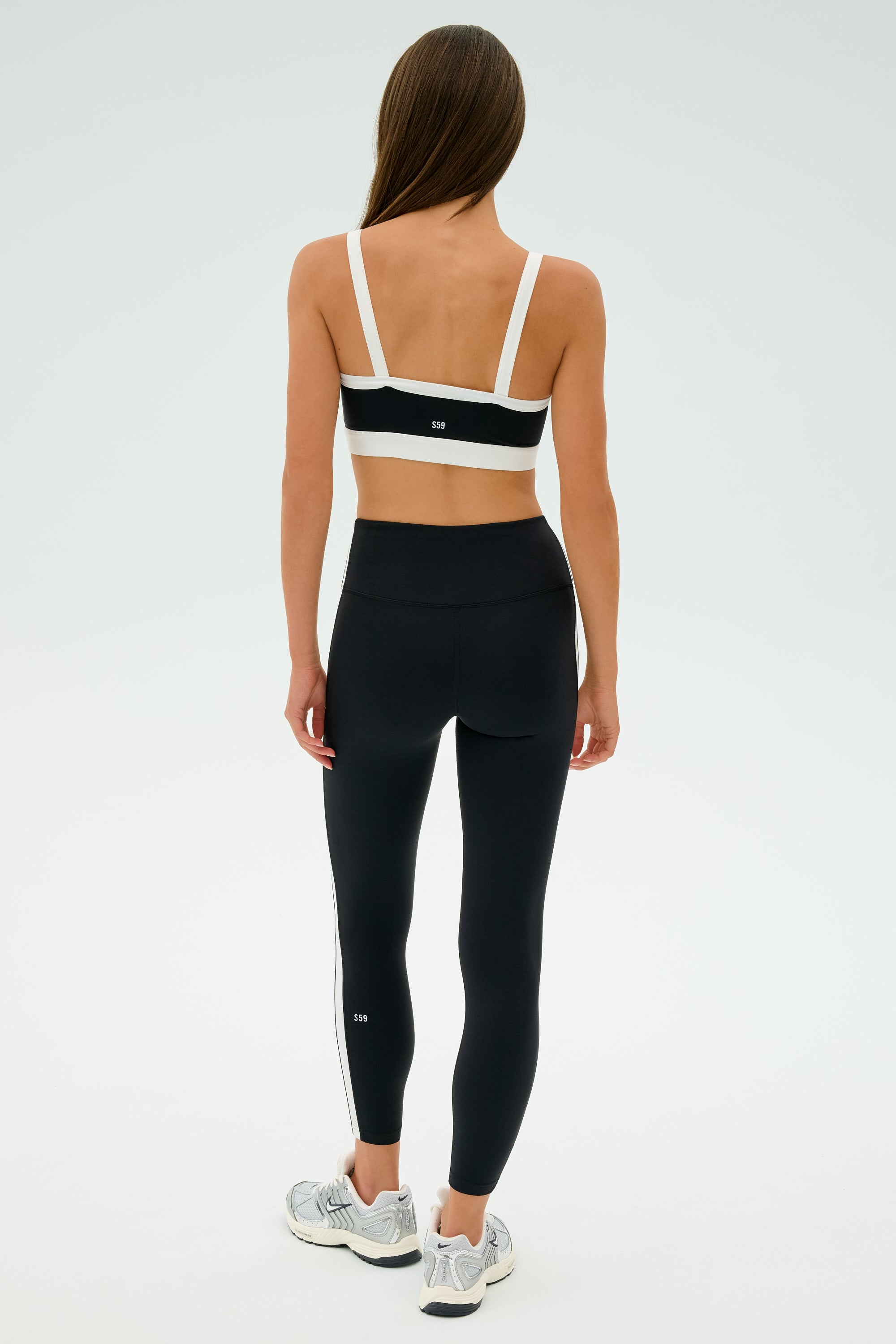 A person dons the SPLITS59 Clare High Waist Rigor 7/8 leggings in black/white, paired with a matching sports bra and white sneakers, facing away on a light background, radiating high performance with each stride.
