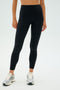A person wearing SPLITS59's Clare High Waist Rigor 7/8 leggings in Black/White paired with white sneakers stands against a light gray background.