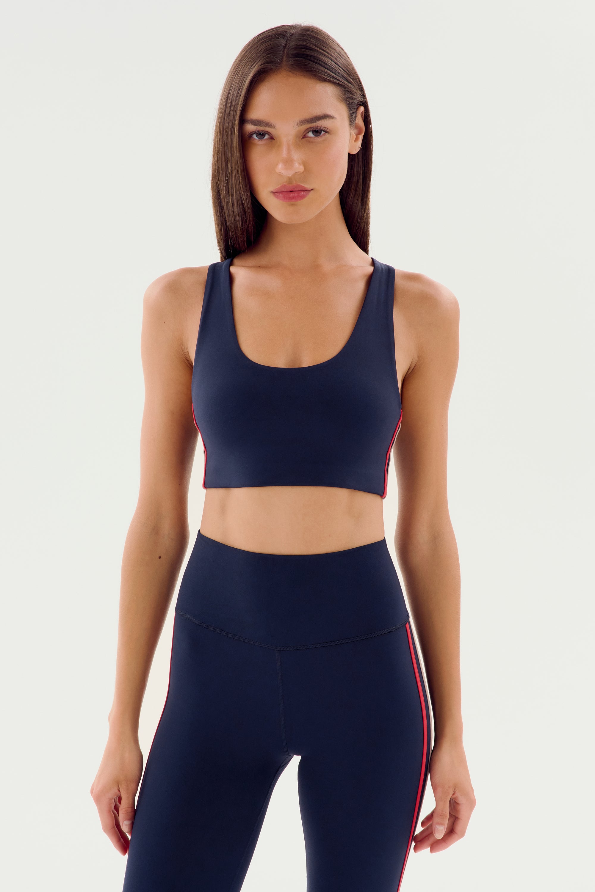 A woman with straight, long hair wearing an Ella Airweight Bra - Indigo/Pirate Red from SPLITS59 and matching navy blue leggings with red stripes from the airweight collection stands against a plain background, perfect for hot yoga sessions.