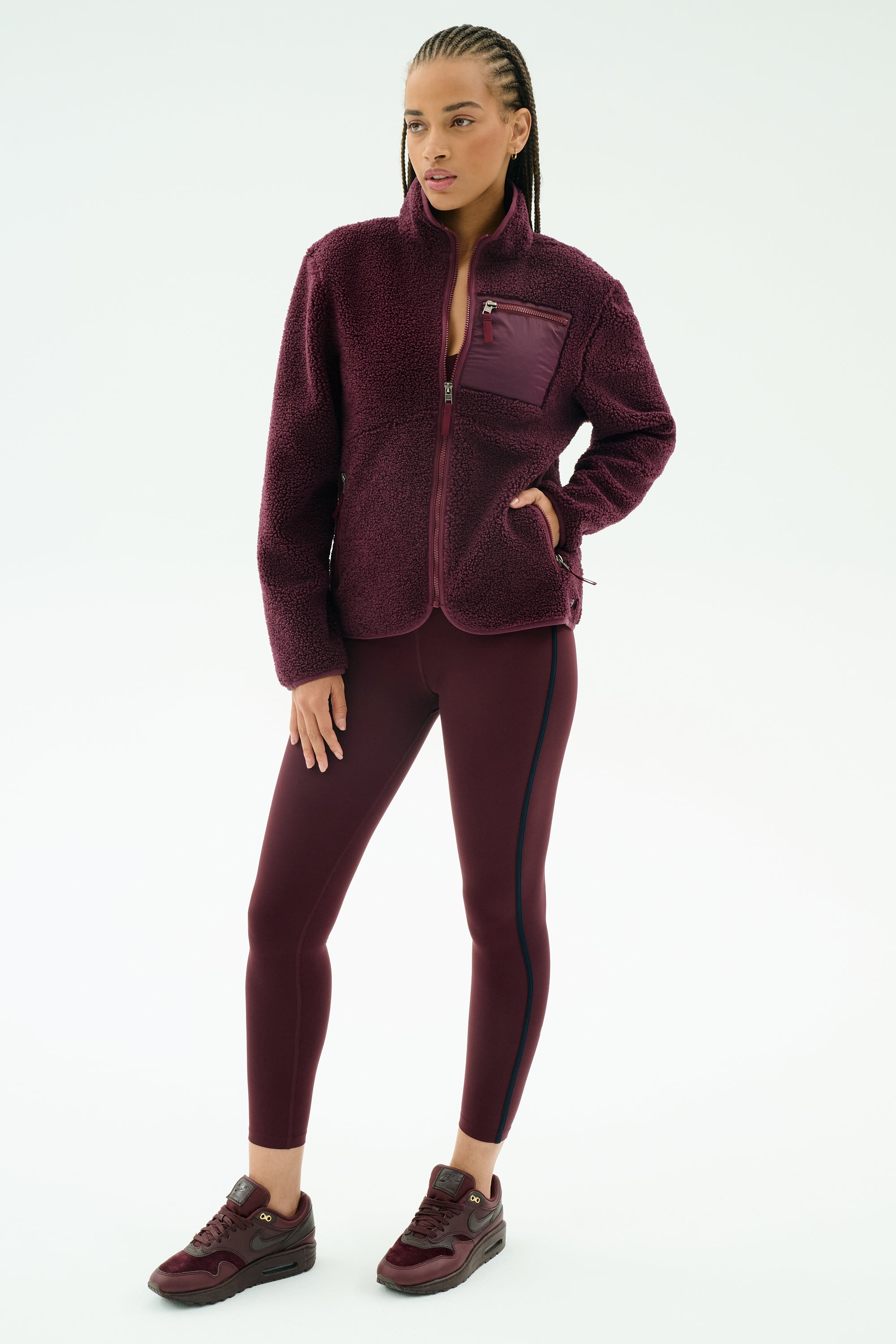 A woman stands against a white background wearing the SPLITS59 Harris Sherpa Jacket in Dark Cherry, paired with matching leggings featuring a black stripe and burgundy sneakers.