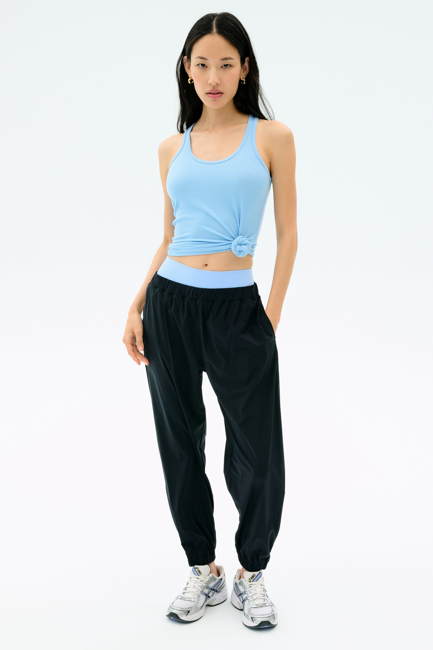 A person stands against a plain background wearing the SPLITS59 Ashby Rib Tank in Skylight, paired with black jogger pants and white sneakers.