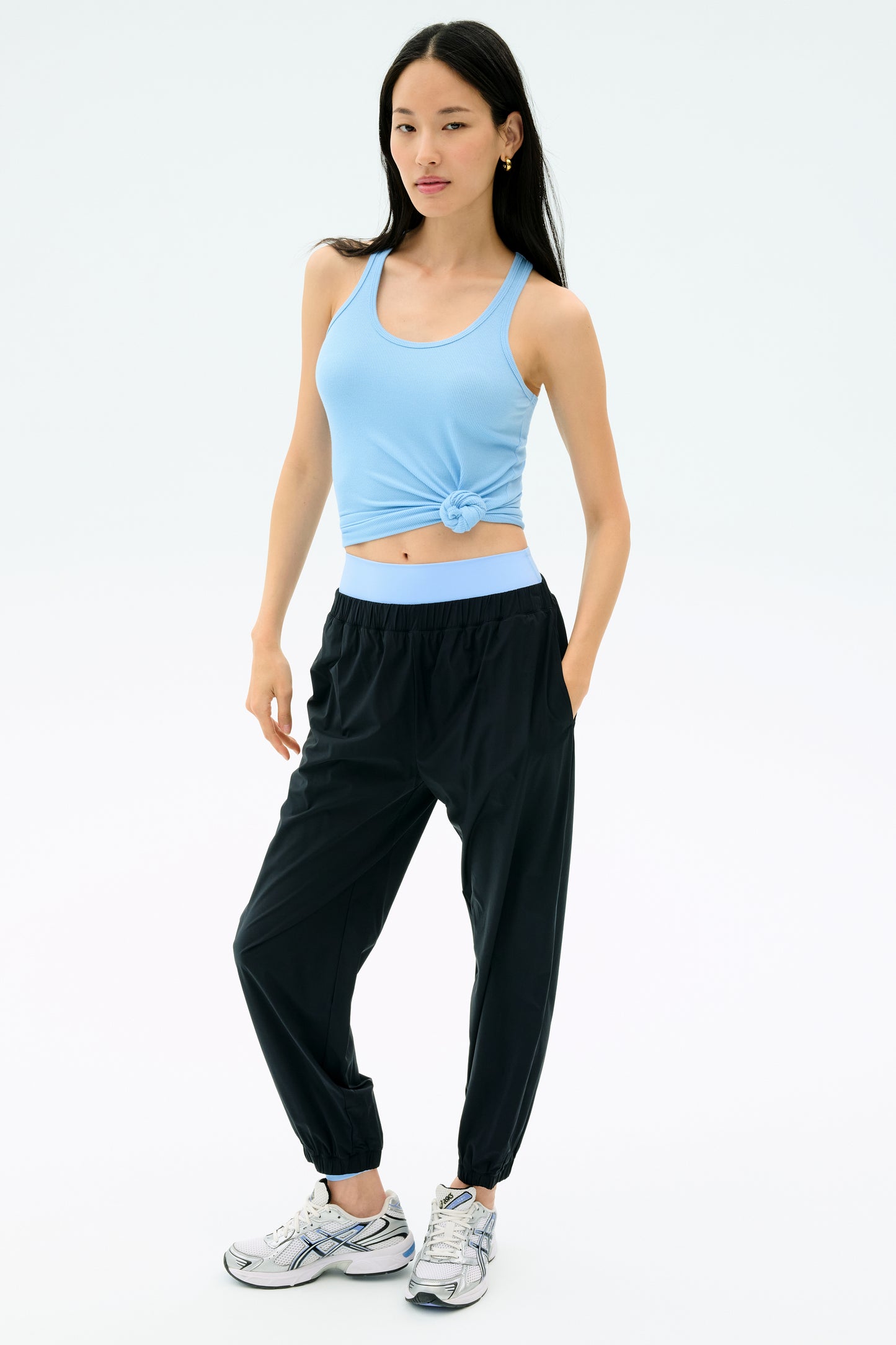 A woman stands against a plain background wearing the SPLITS59 Ashby Rib Tank in Skylight, paired with black joggers and white sneakers, her left hand in her pocket.