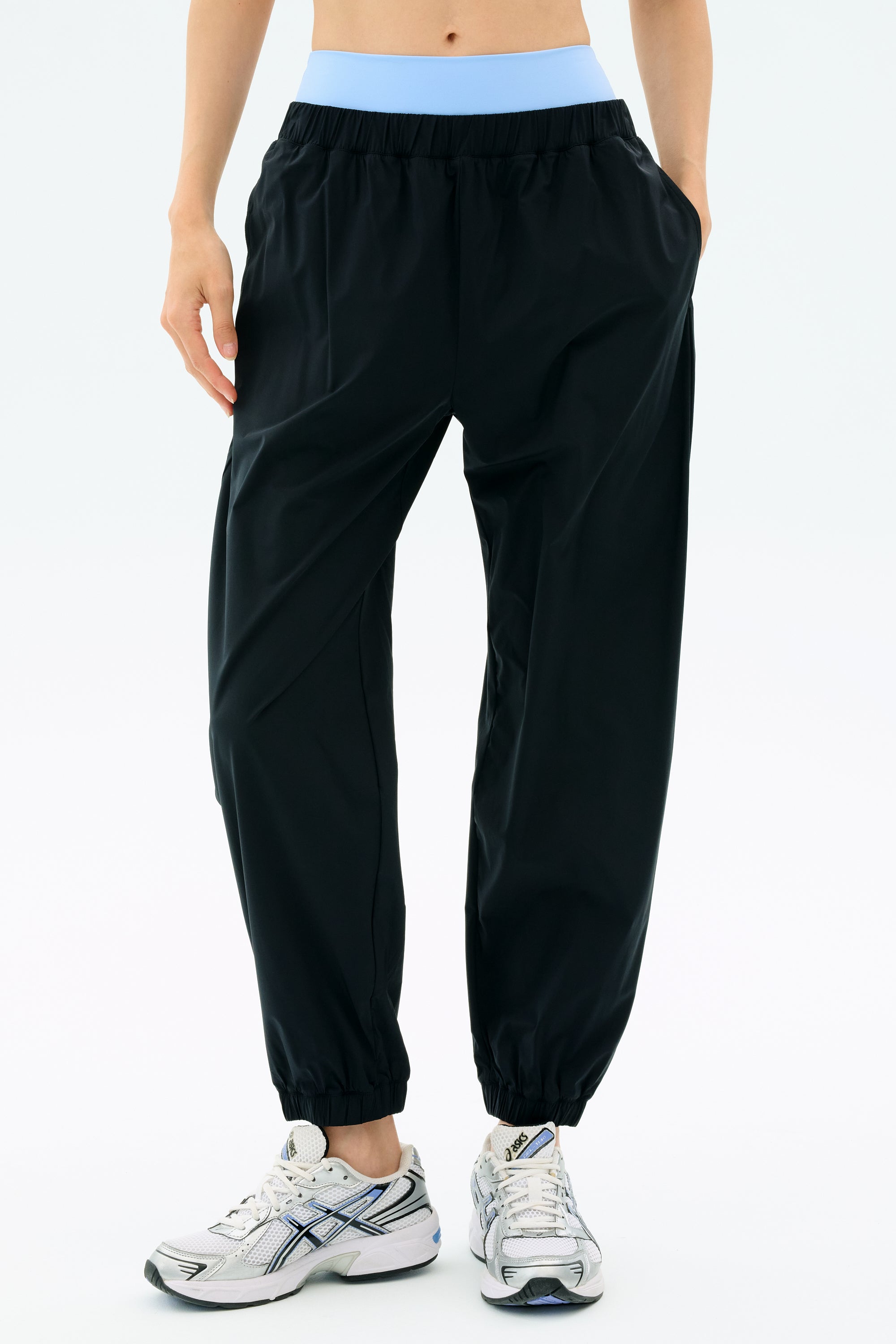 A person wearing the SPLITS59 Megan Woven Nylon Jogger in black with a blue waistband and white athletic shoes stands against a light background.