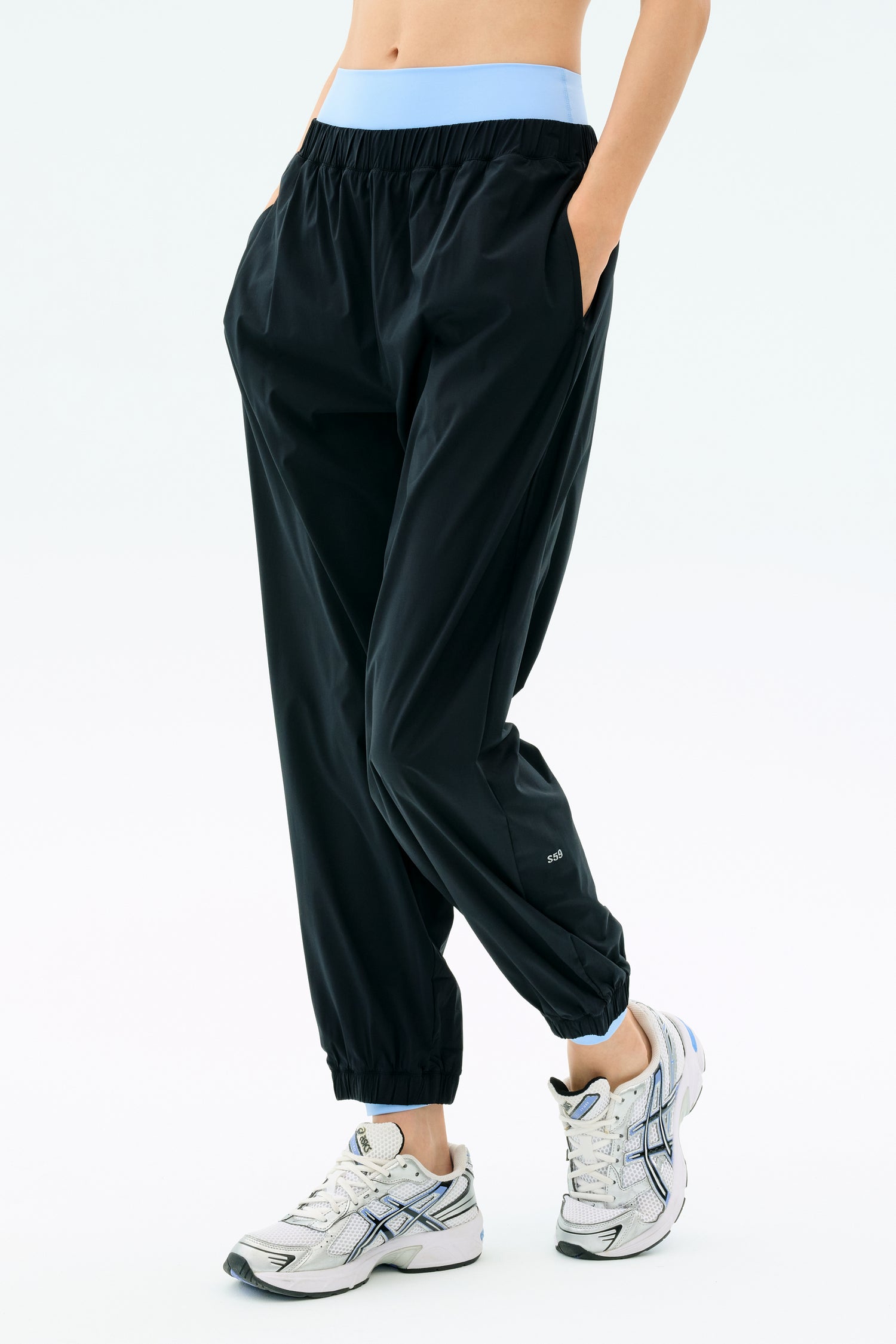 A person wearing SPLITS59 Megan Woven Nylon Jogger in black, featuring a light blue spandex waistband, with white athletic shoes and hands in pockets, stands against a plain light background.