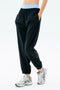 A person wearing SPLITS59 Megan Woven Nylon Jogger in black, featuring a light blue spandex waistband, with white athletic shoes and hands in pockets, stands against a plain light background.