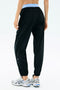 Someone wearing SPLITS59 Megan Woven Nylon Jogger in black, featuring a light blue waistband, paired with white athletic shoes, is shown from the back against a plain background.