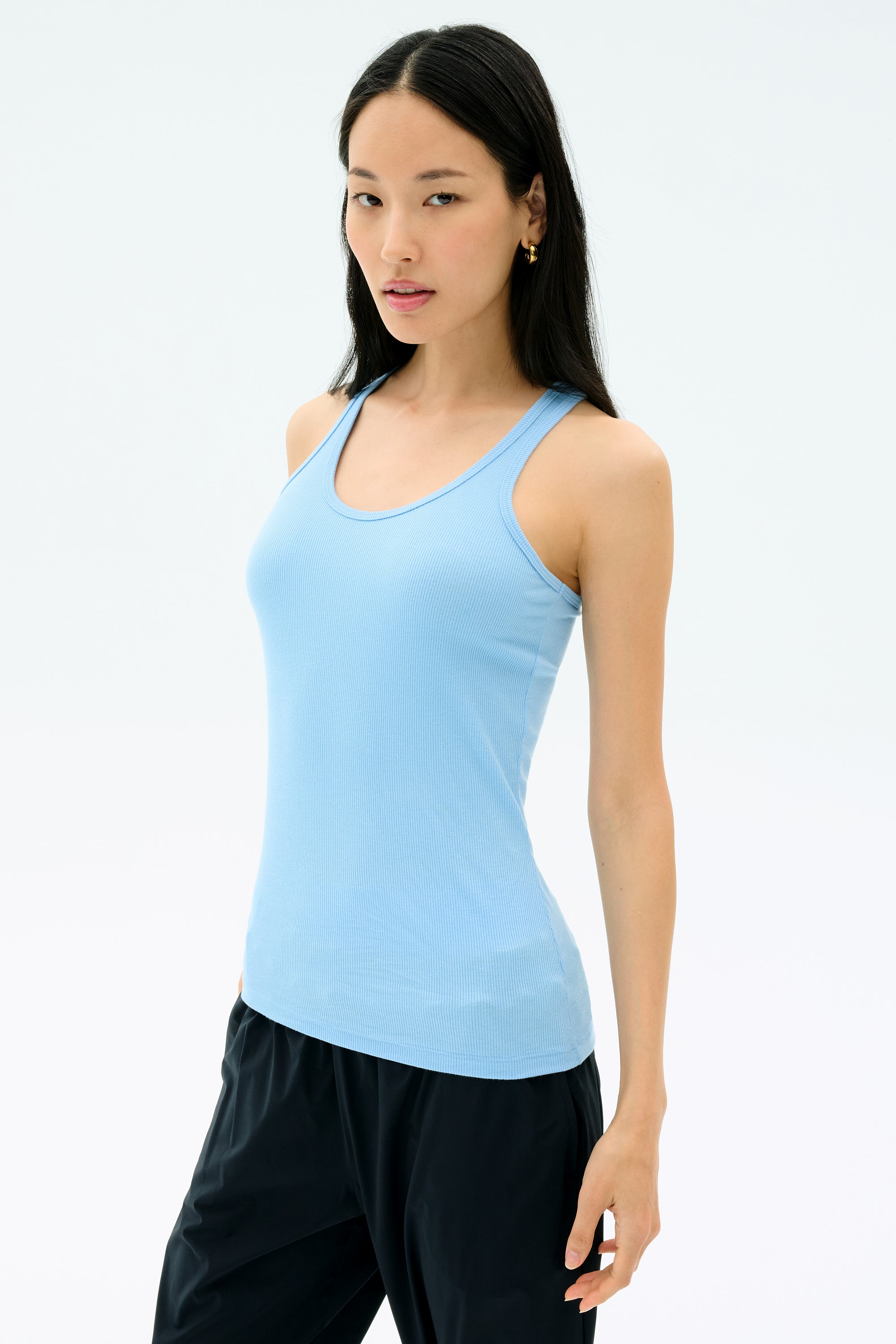 A woman wears the SPLITS59 Ashby Rib Tank in Skylight and black pants, crafted from soft baby rib fabric, standing against a plain white background.