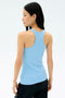 A woman with long black hair wears the SPLITS59 Ashby Rib Tank in Skylight and black pants, standing with her back to a plain light background—perfect attire for gym sessions or yoga.