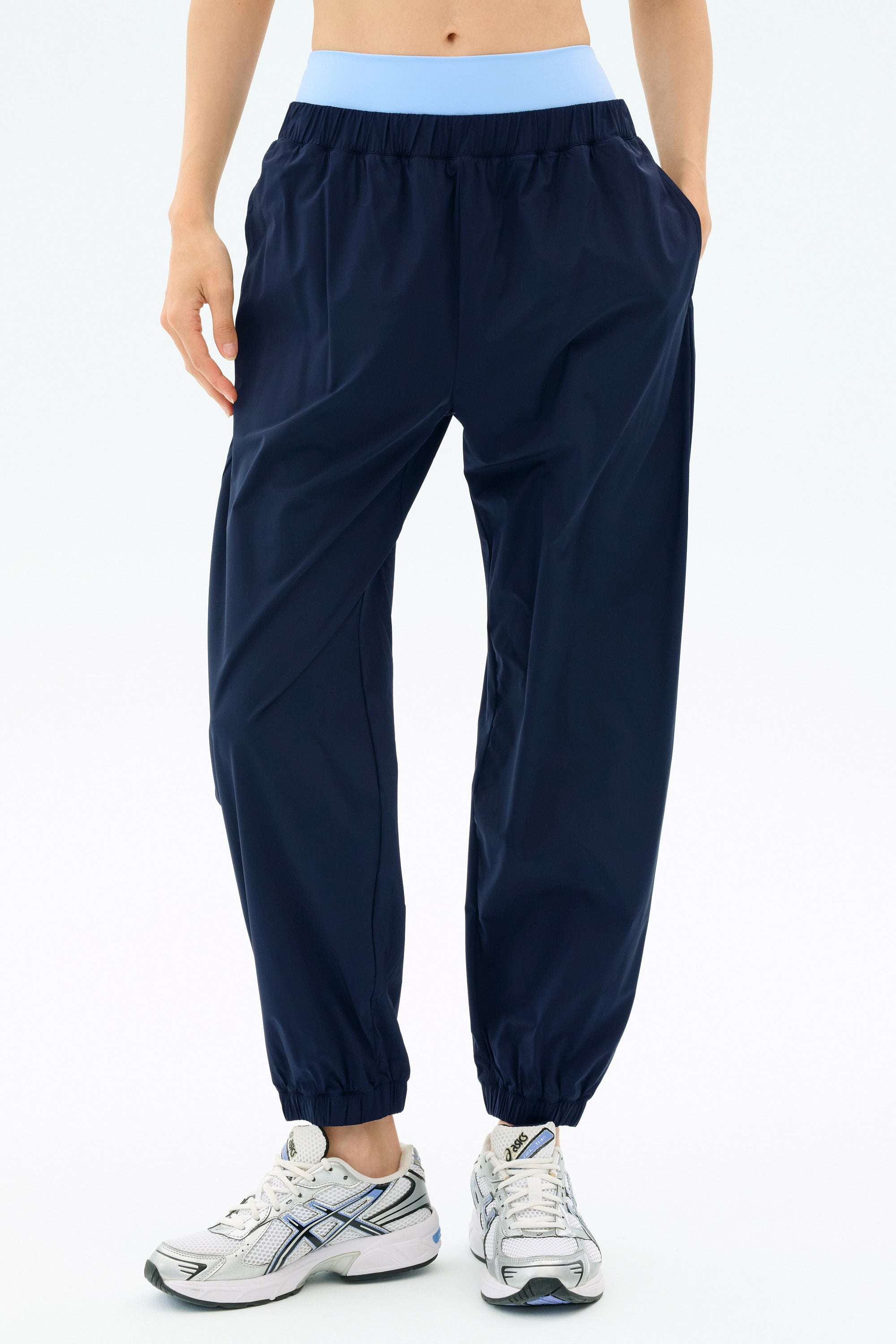 Model wearing SPLITS59 Megan Woven Nylon Jogger in Indigo, featuring elastic cuffs and a light blue waistband, paired with white sneakers and hands in pockets, standing against a plain background.