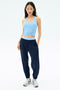 A woman wearing a light blue knotted tank top, SPLITS59 Megan Woven Nylon Jogger in Indigo, and white sneakers stands against a plain white background.
